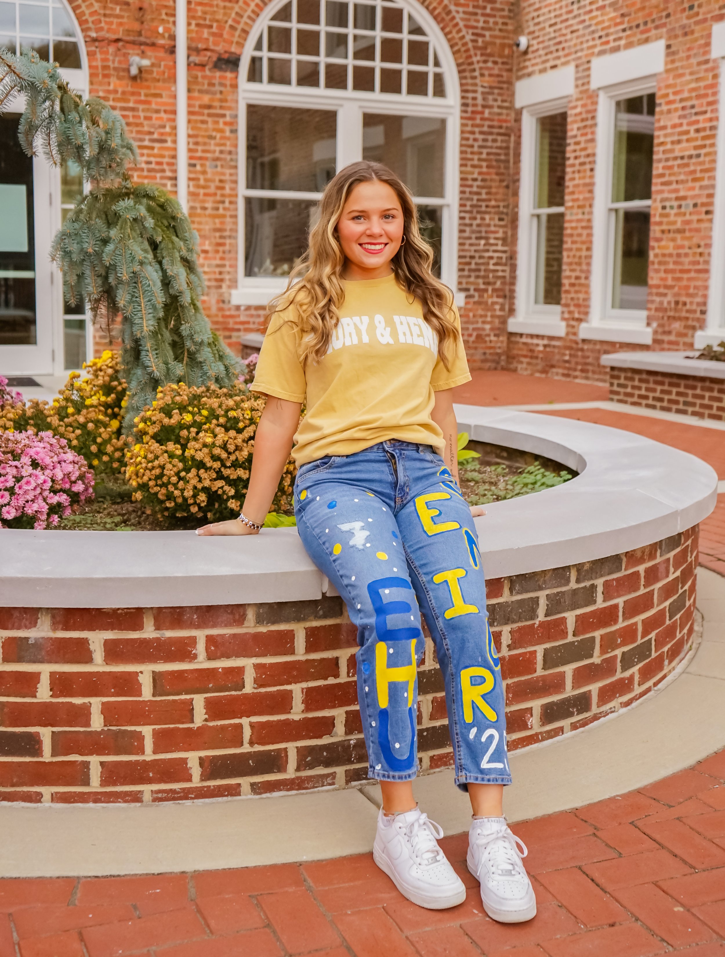 Young woman sitting on a brick planter in front of a red brick building with large arched windows, wearing a yellow T-shirt and decorated jeans spelling out 'EHTER' with colorful letters.