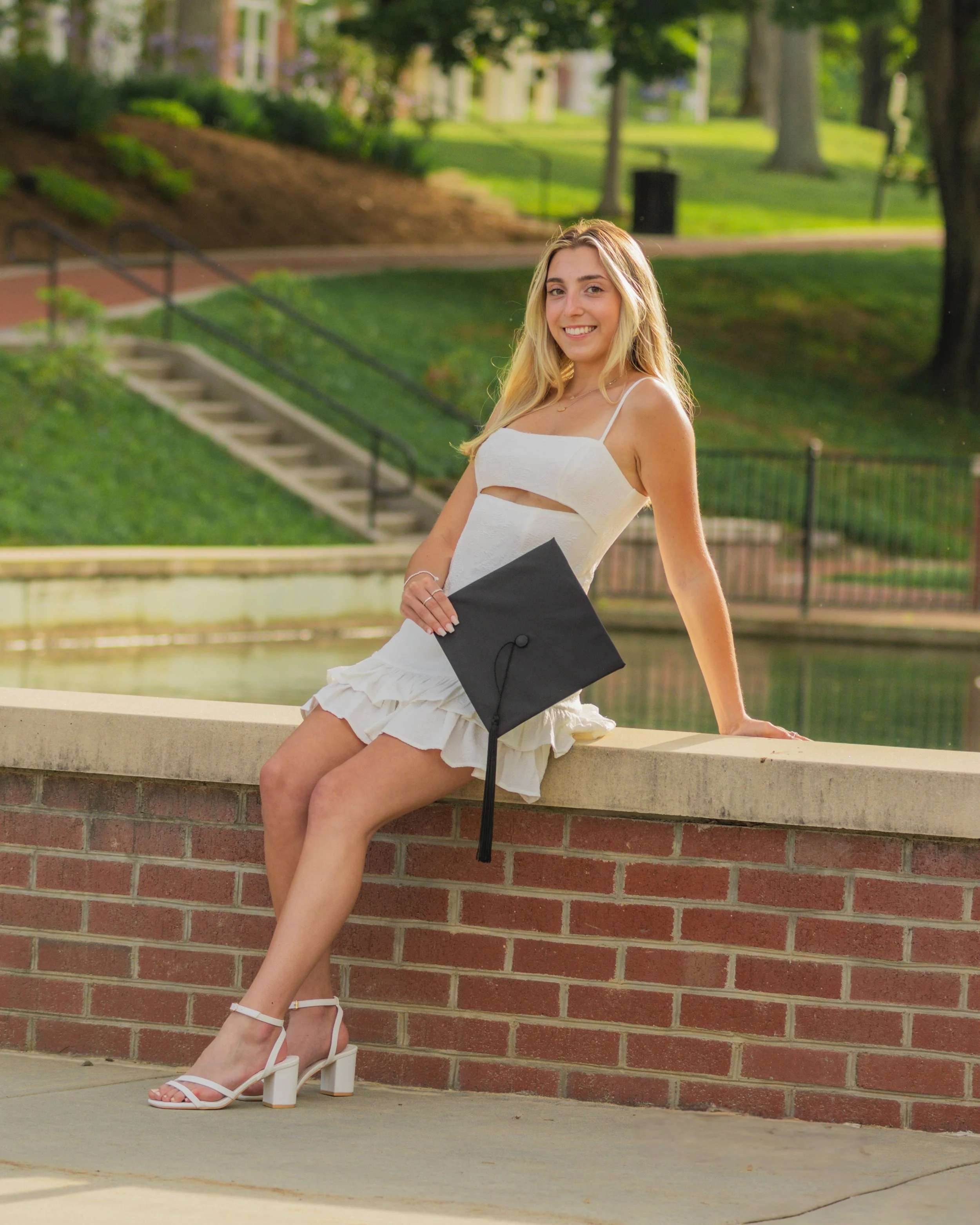 Young woman in a white dress with spaghetti straps sitting on a brick ledge, holding a black graduation cap, smiling outdoors near a pond in a park.
