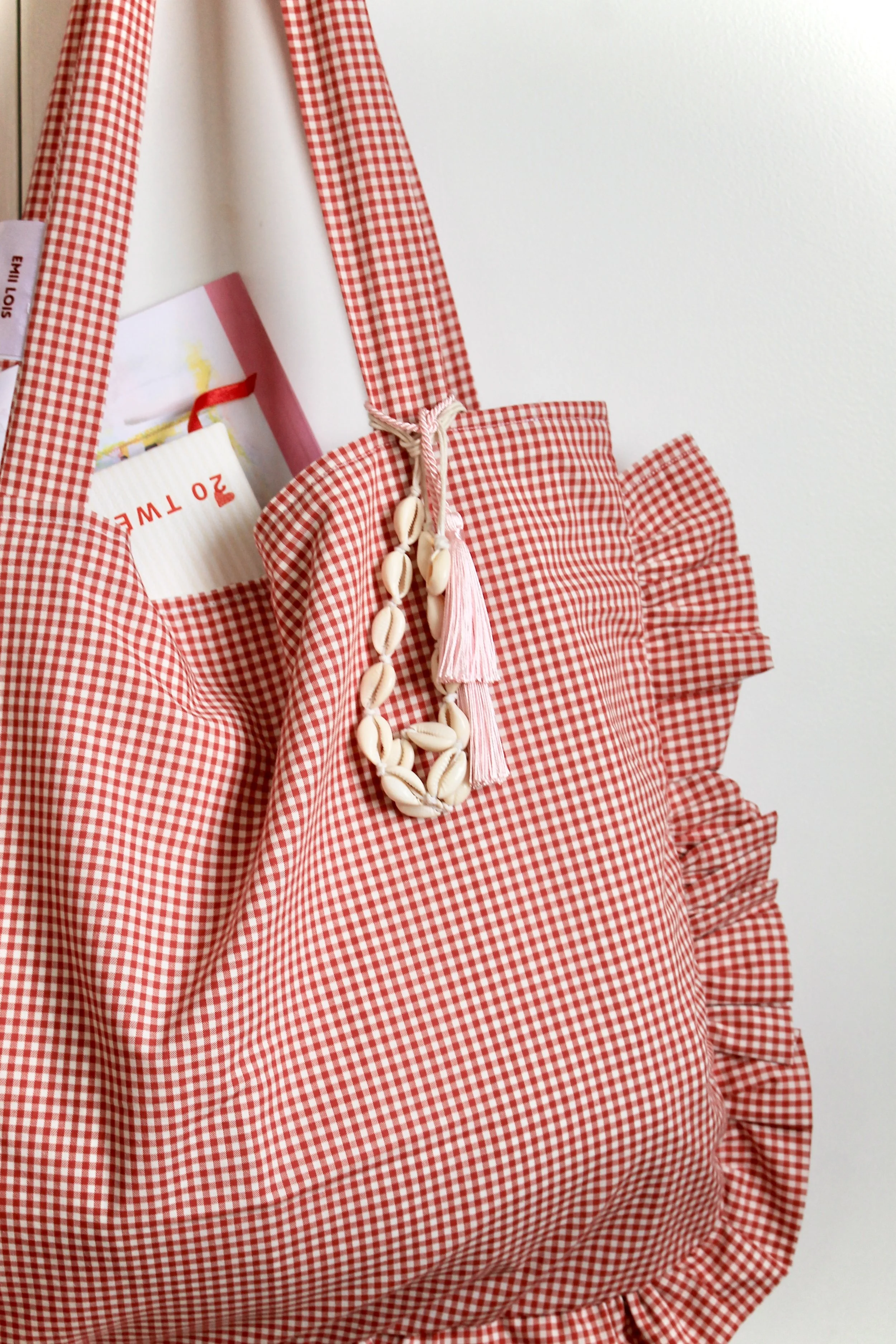Red gingham tote bag with ruffles and a shell necklace, containing a book with a pink and white cover.