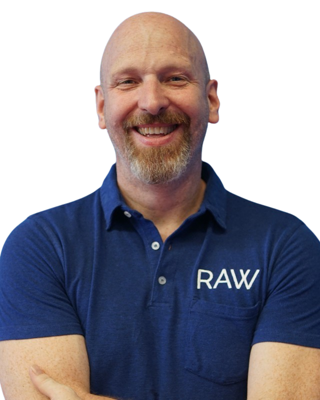 A smiling man with a beard and bald head wearing a blue polo shirt with "RAW" embroidered on it, standing with arms crossed.