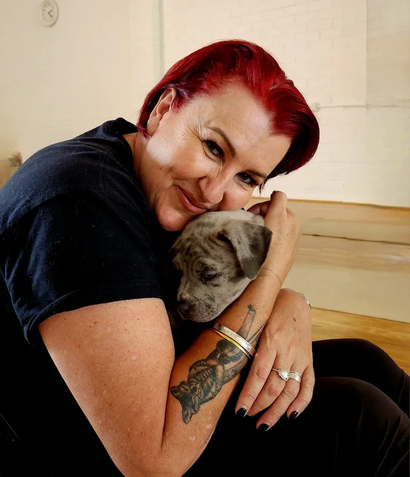 A woman with red hair holding a small, gray puppy close to her face, smiling and gently cradling the puppy's head.