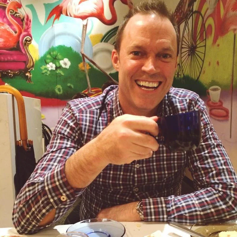 A smiling man wearing a plaid shirt sitting at a table, holding a dark blue mug, with colorful graffiti art mural in the background.