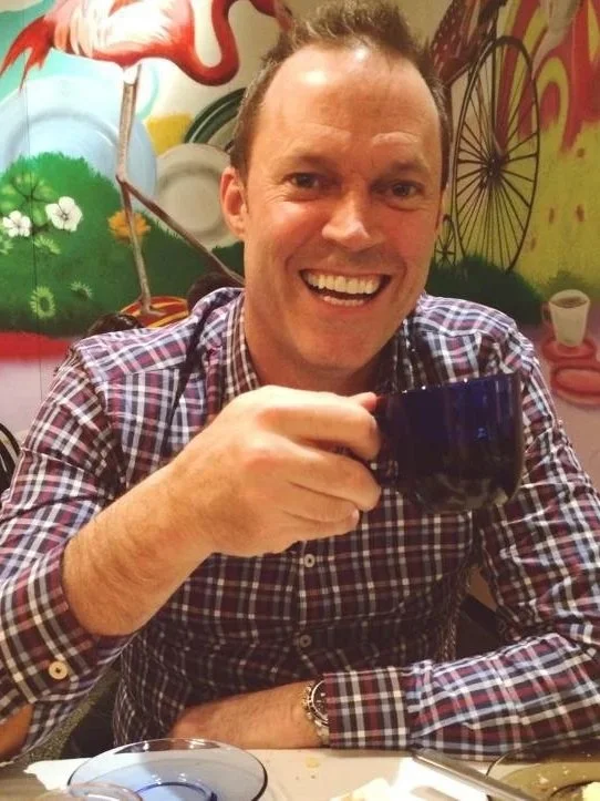 A smiling man wearing a plaid shirt sitting at a table, holding a dark blue mug, with colorful graffiti art mural in the background.