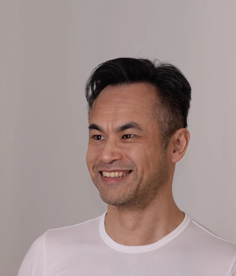 Portrait of a smiling man with dark hair slicked back, wearing a white shirt, standing against a plain gray background.