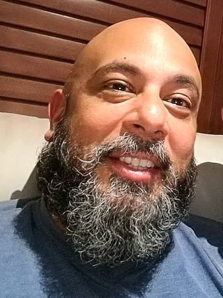 A smiling man with a bald head and a full gray beard, wearing a dark shirt, sitting indoors with a wooden wall in the background.