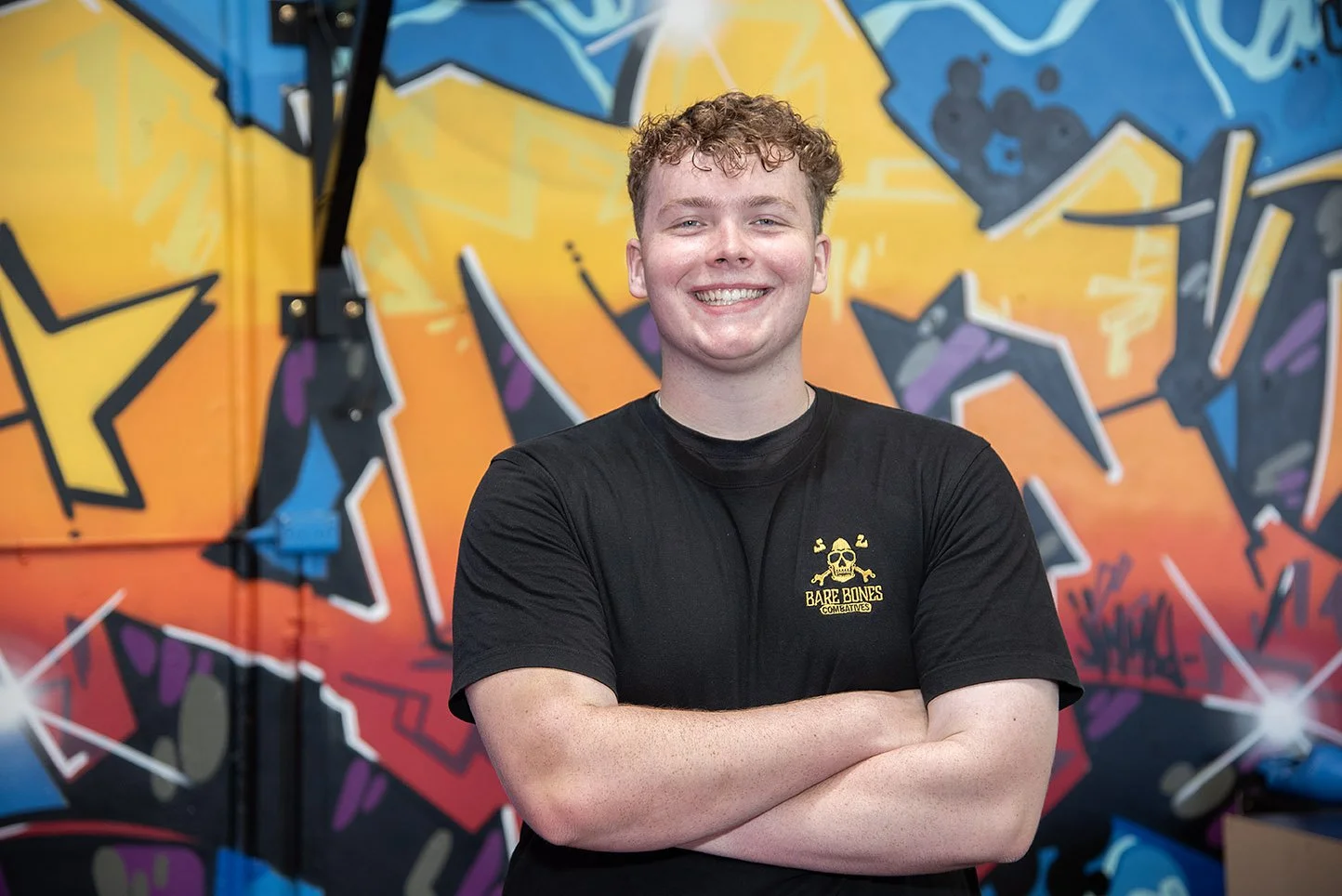 Geordie Armstrong, kickboxing and fitness coach at Raw Life Australia in Rowville, standing in front of a colourful graffiti wall wearing a BareBones Combatives T-shirt.
