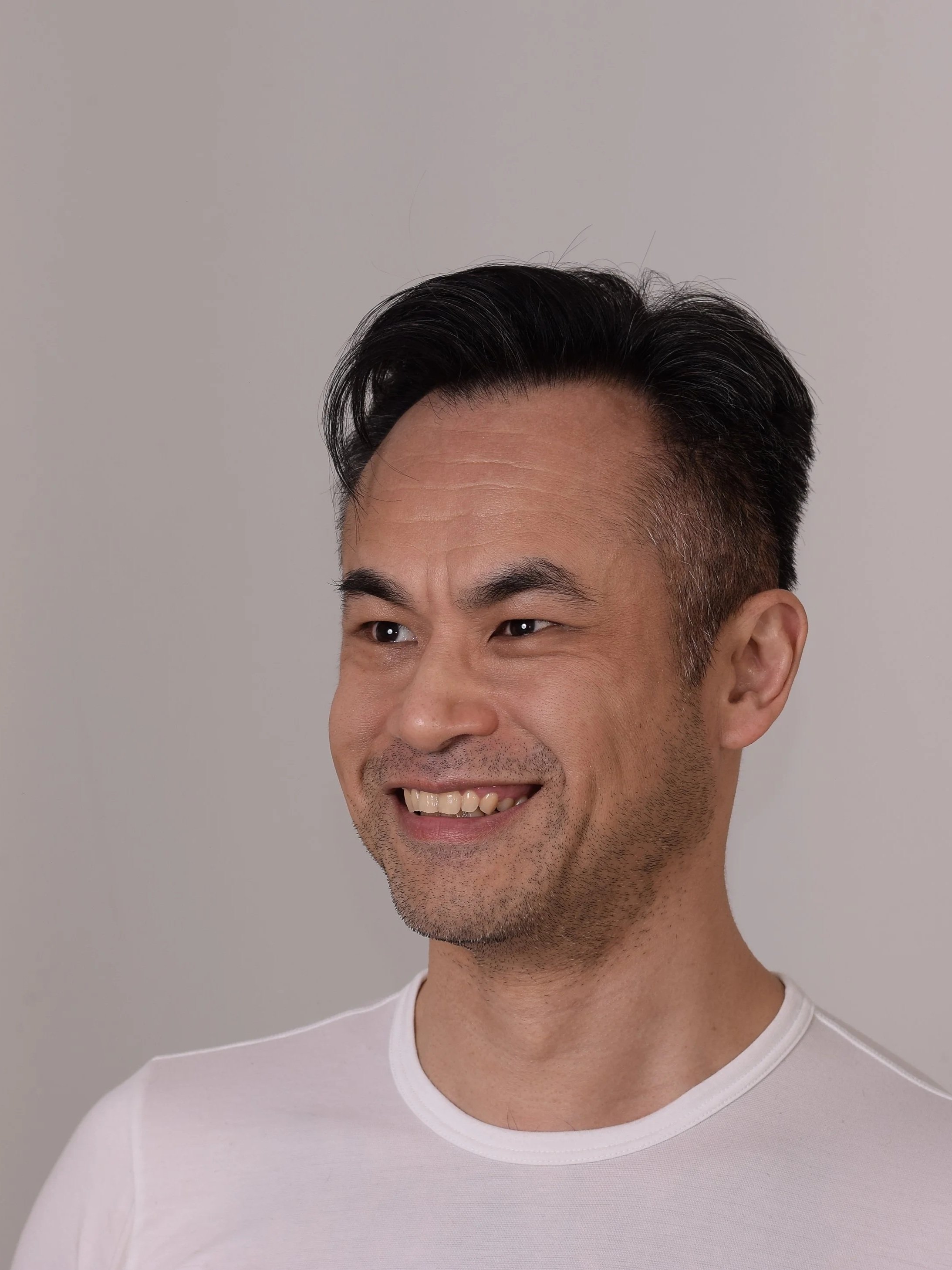Portrait of a smiling man with dark hair slicked back, wearing a white shirt, standing against a plain gray background.