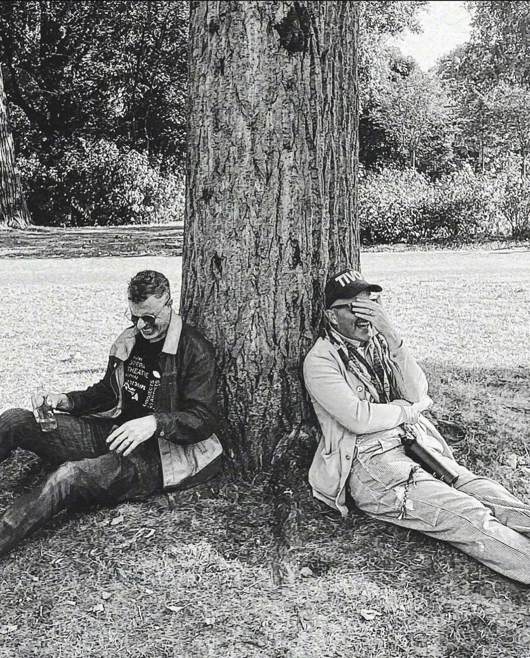 Matt Ray and Taylor Mac sitting on the ground leaning against a large tree trunk, Matt with sunglasses and holding a drink, Taylor with a cap and binoculars, covering face with hand, in a park or outdoor setting.