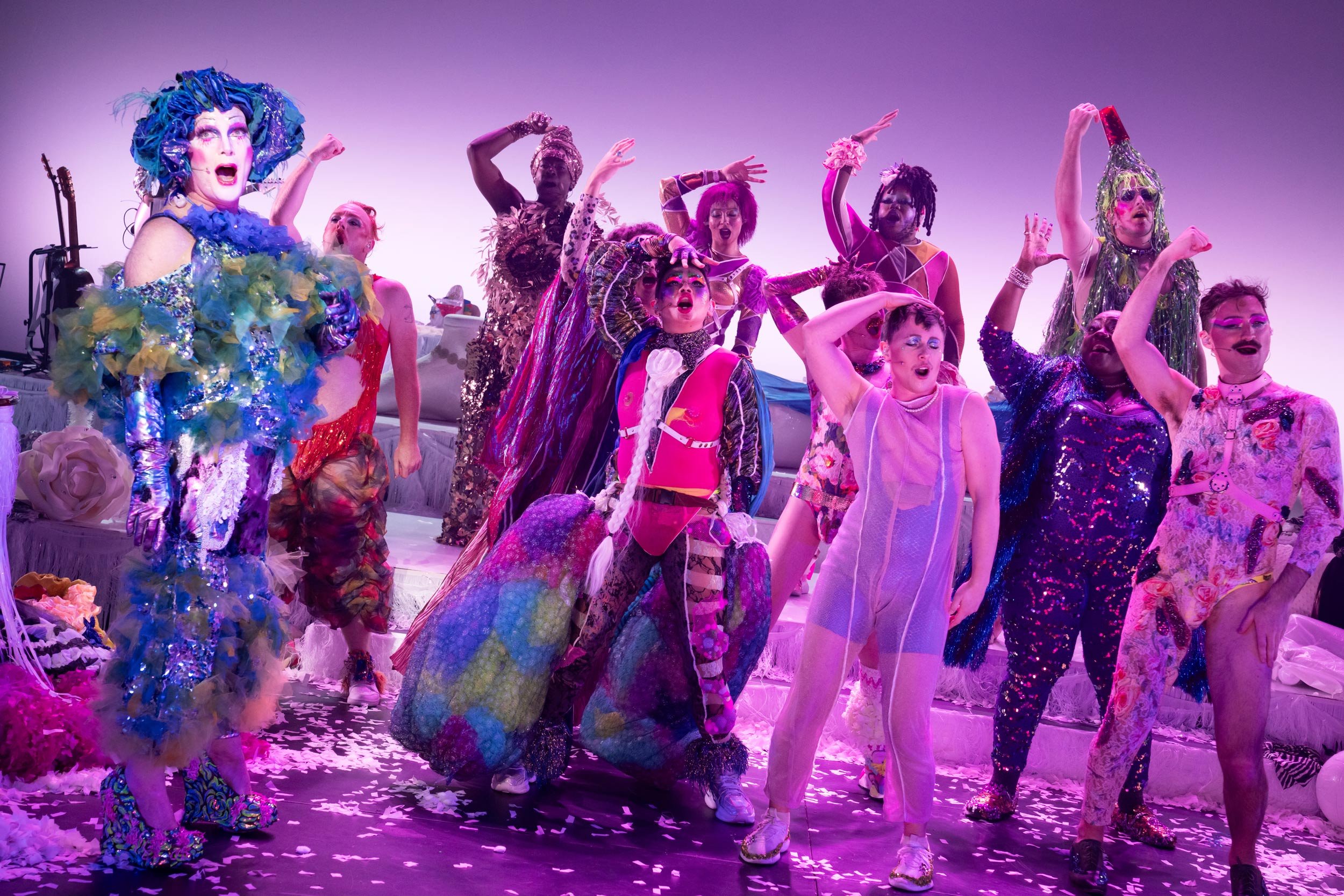 Colorful performers in vibrant costumes on stage during a lively drag show, with a purple-lit backdrop and scattered confetti.