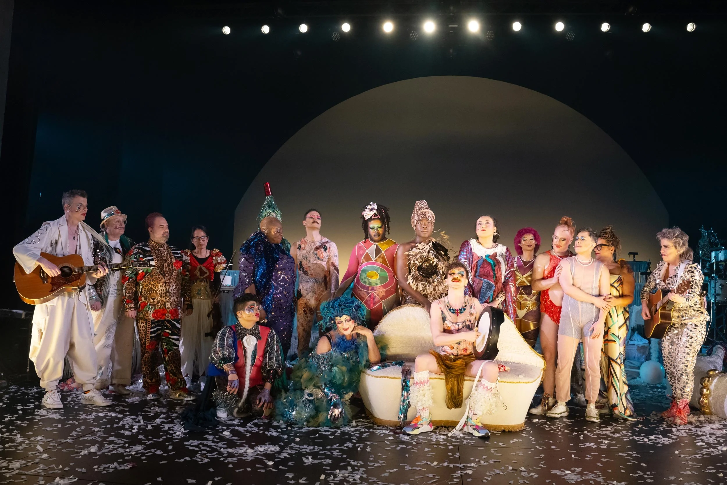 Group of performers on stage dressed in vibrant, colorful costumes with makeup and accessories, standing amid confetti and decorative props.