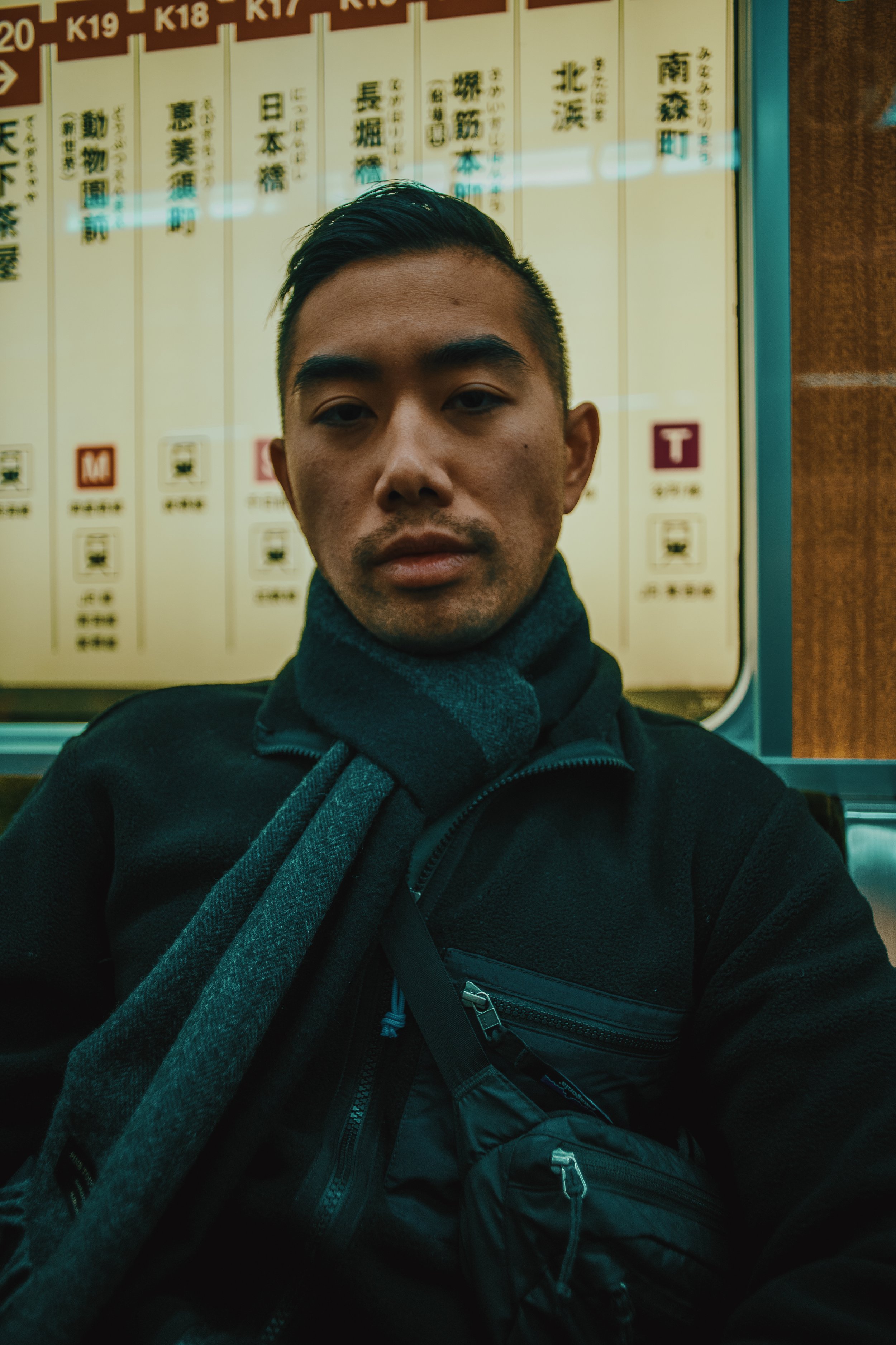 A young man with short dark hair and a serious expression sitting in a transportation vehicle, with a large station or route map with Japanese characters behind him.