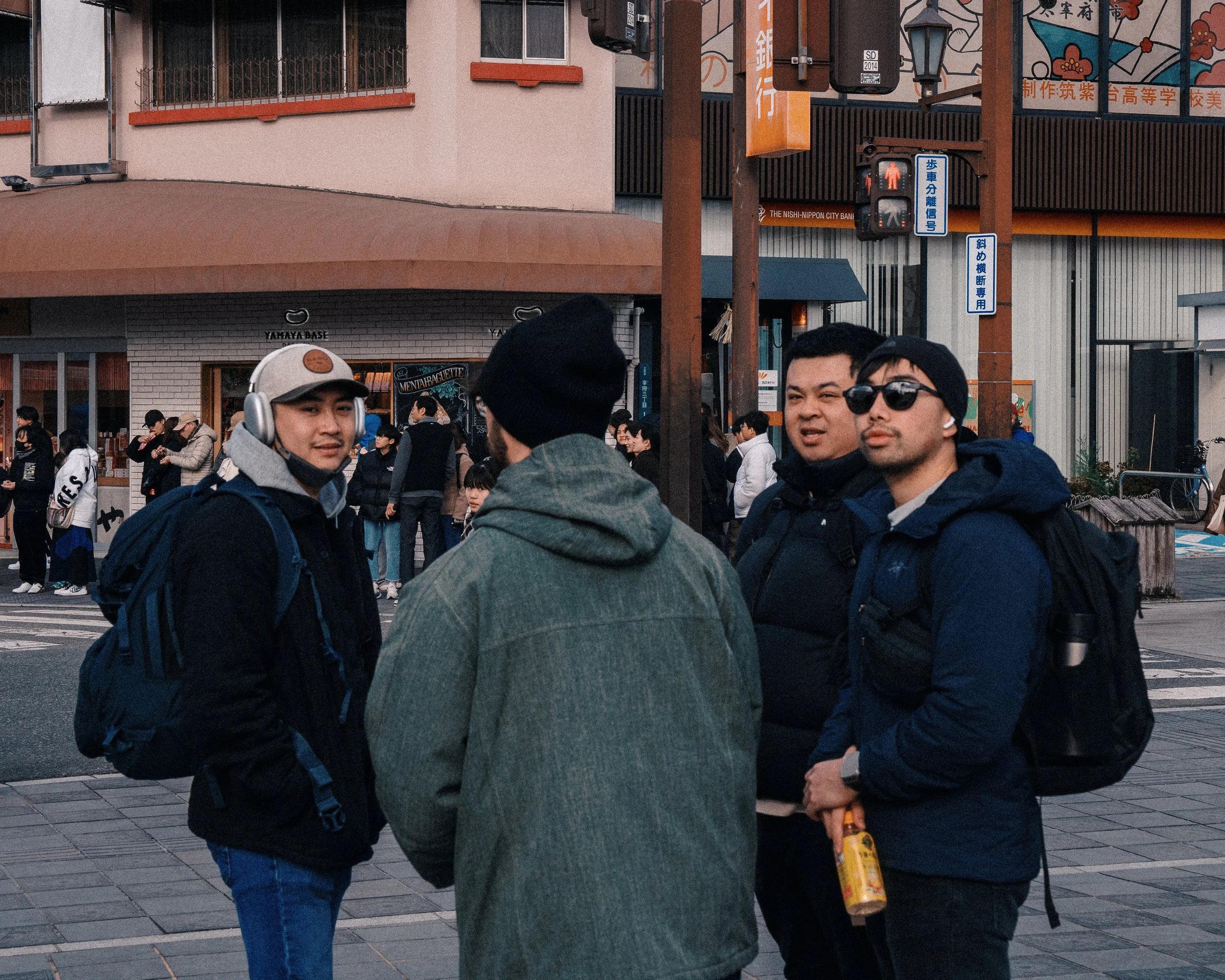 Four young men standing on a city street corner talking, with a crowd and buildings in the background.