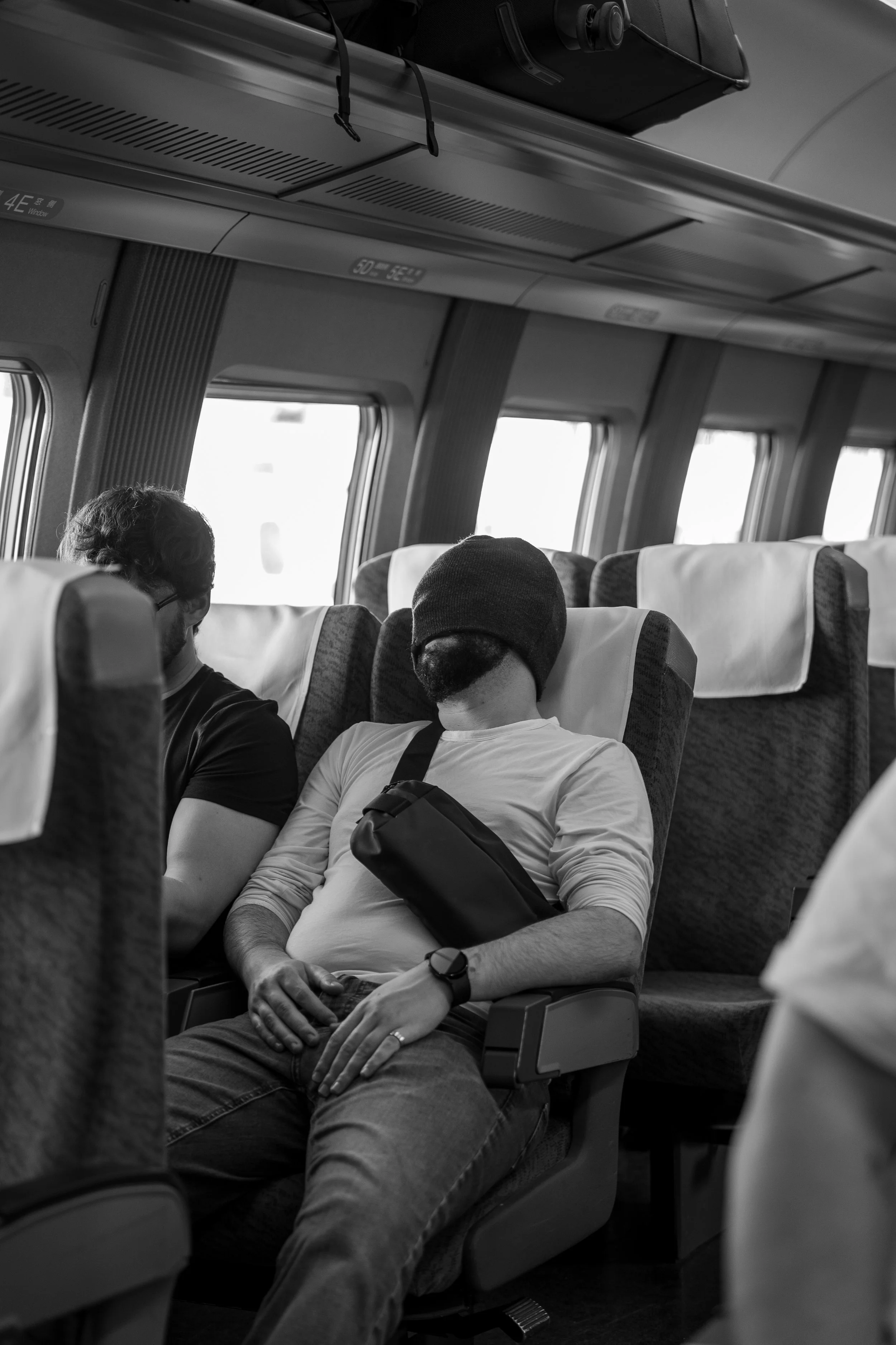 A man with a beanie sleeping on an airplane seat while another passenger is seated next to him.