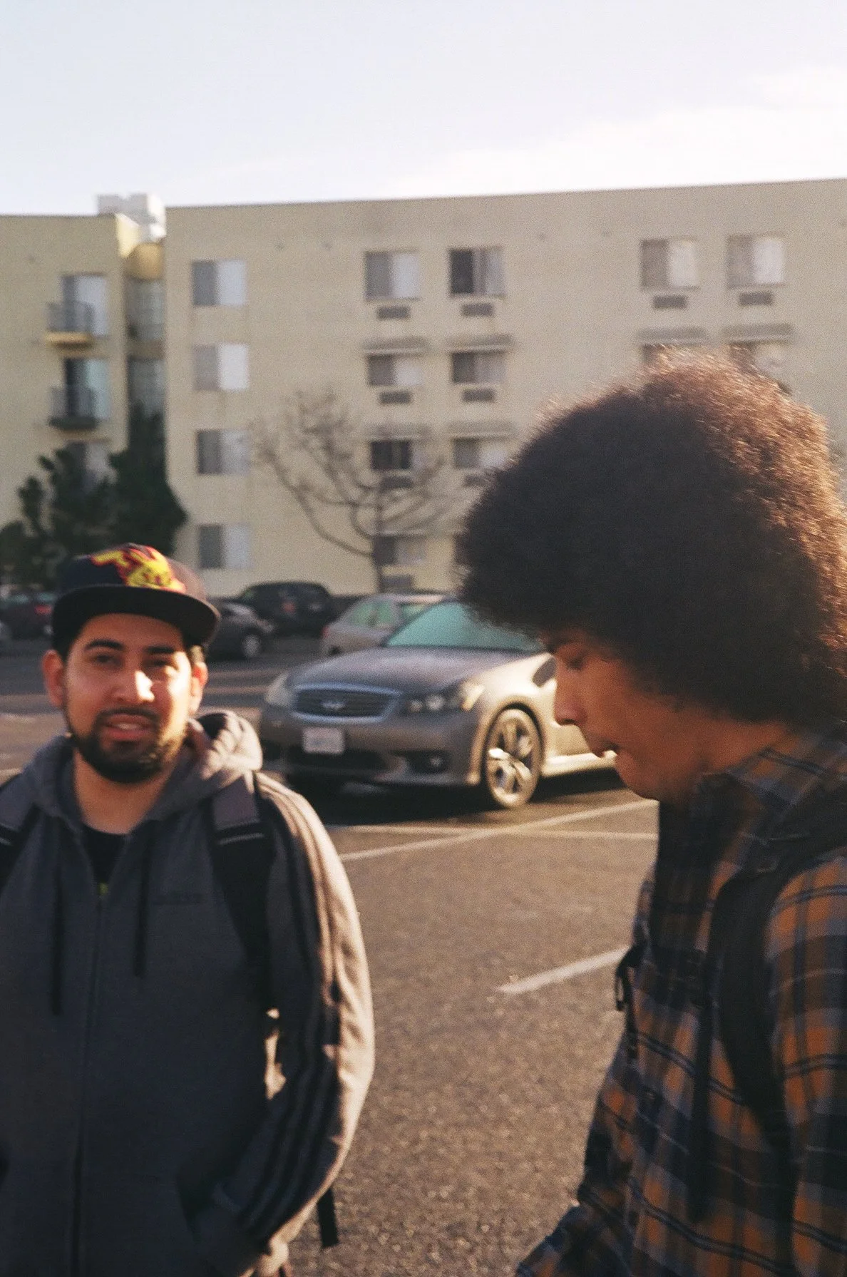 Two men standing outdoors near parked cars with a multi-story apartment building in the background. One man is looking at the camera, wearing a dark jacket and a cap, while the other man is looking down, with a plaid shirt and large afro hairstyle.