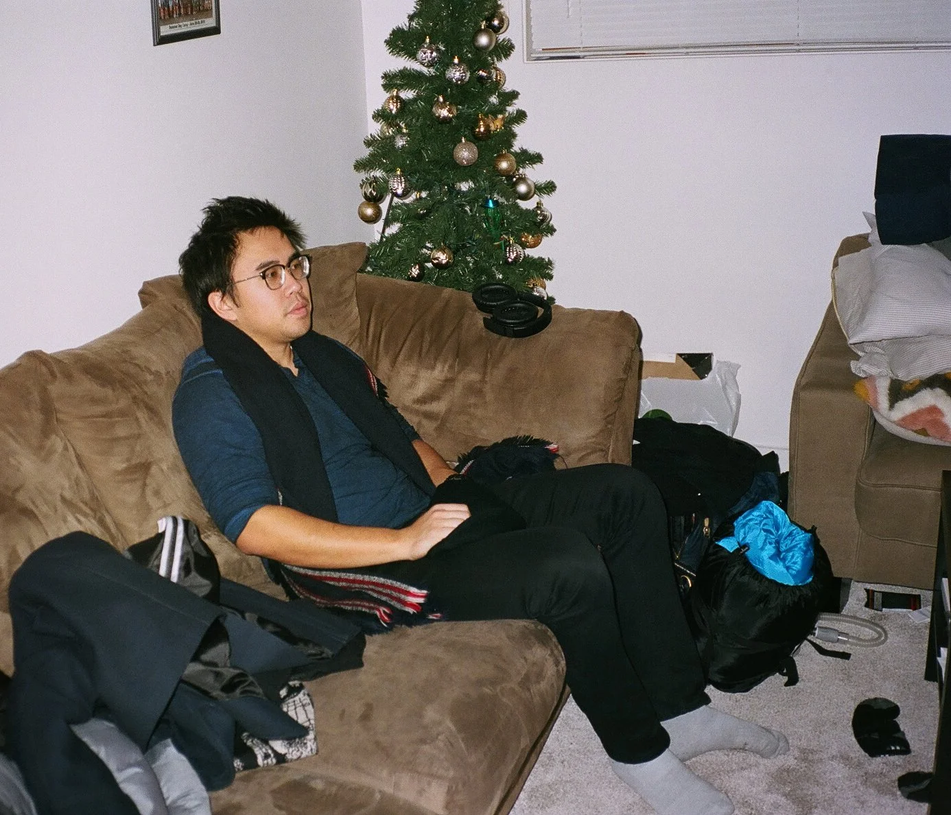 A man sitting on a brown couch with a Christmas tree in the background, surrounded by clothes and bags.