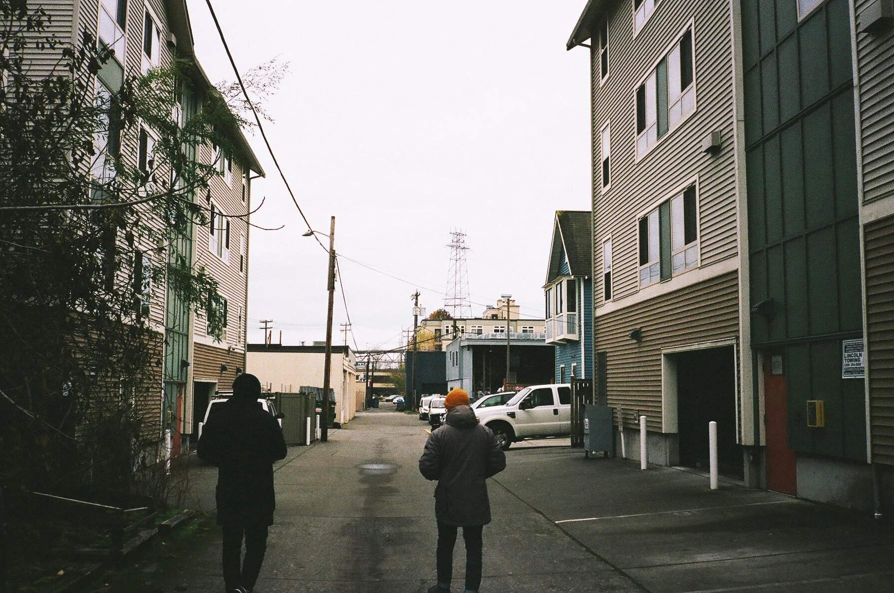 Two people walking in an alleyway surrounded by multi-story buildings, parked cars, and visible power lines on a cloudy day.
