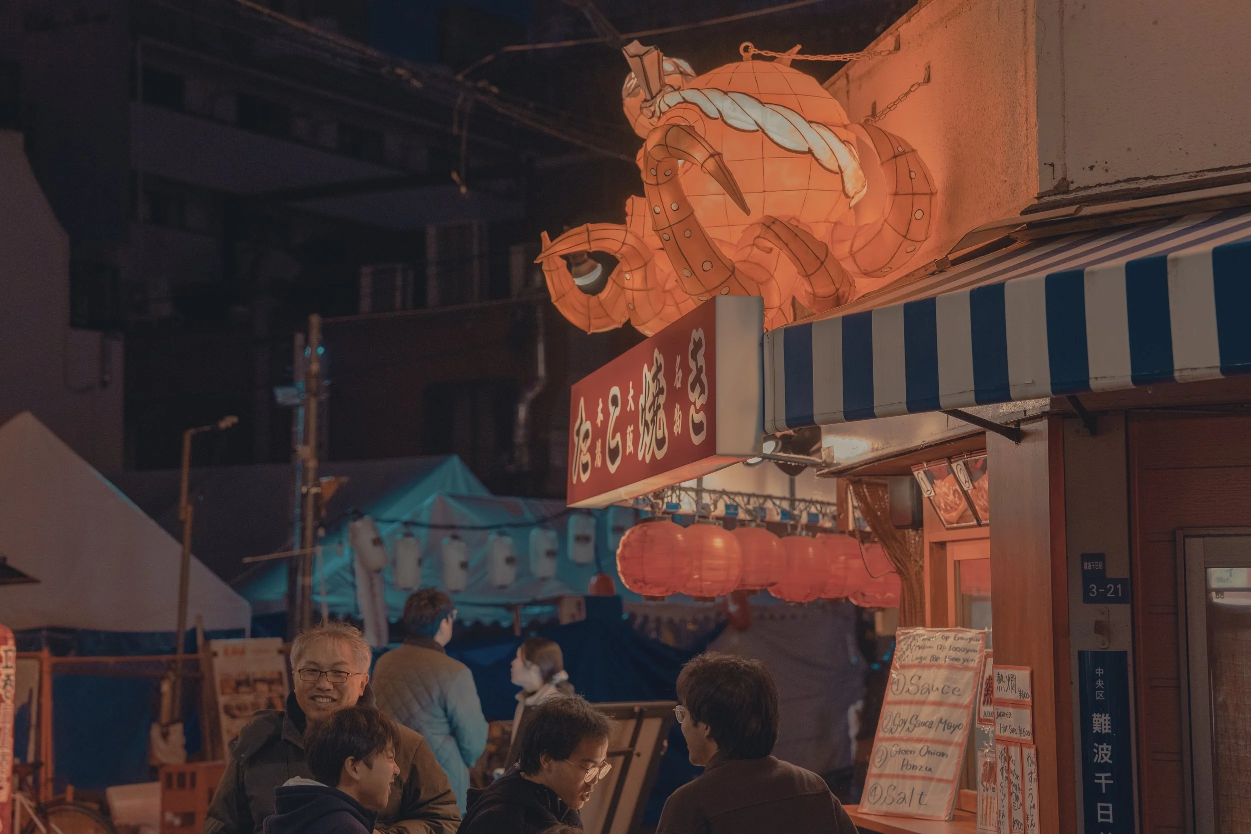 Night scene at a street food stall with illuminated orange lanterns, a large illuminated crab lantern on top, Japanese signs, and people gathered, chatting and enjoying food.