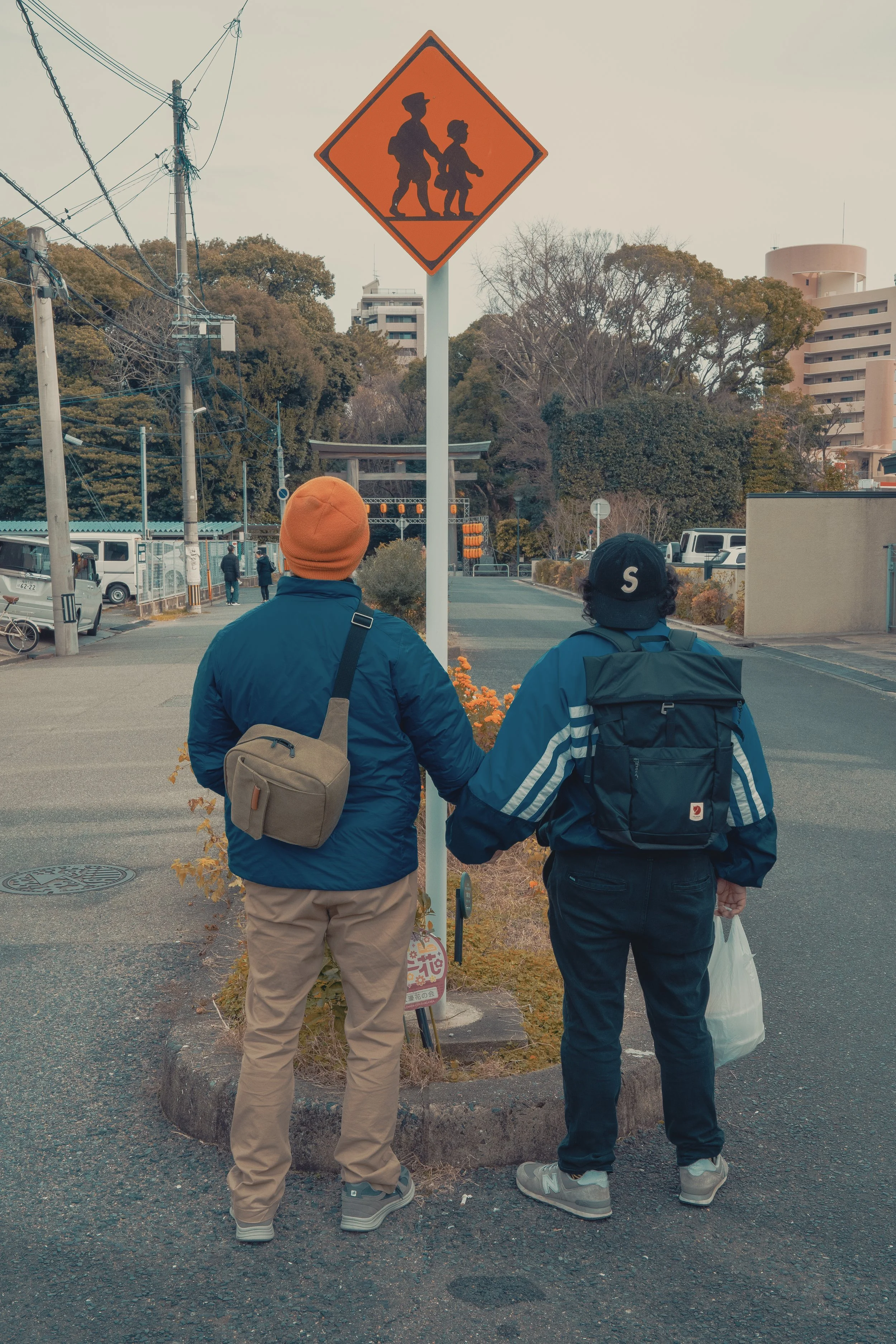 Two people holding hands and standing under a traffic sign showing children crossing, on a street with trees and buildings in the background.
