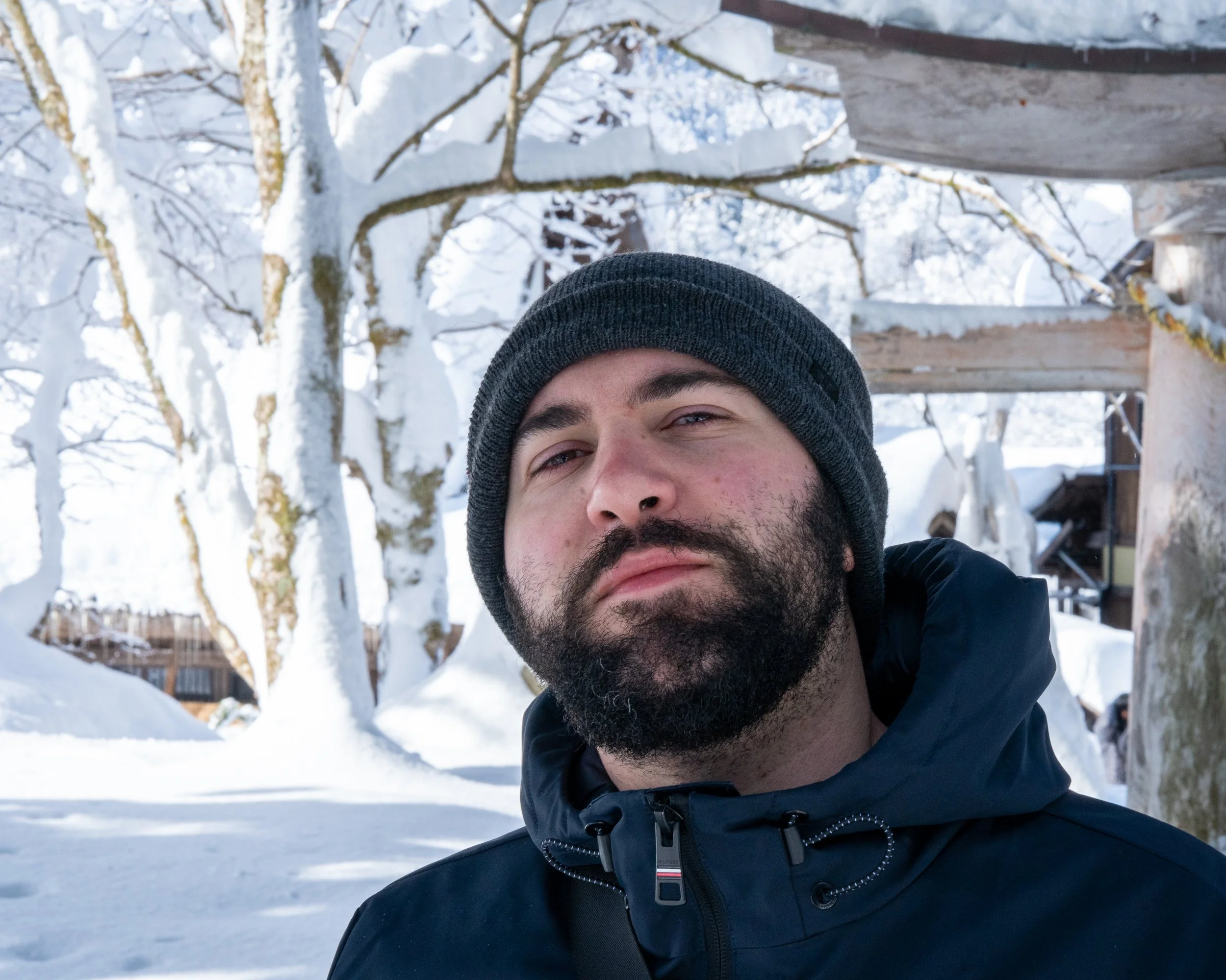 A man with a beard wearing a dark beanie and winter jacket standing outside in a snow-covered landscape with trees and structures in the background.