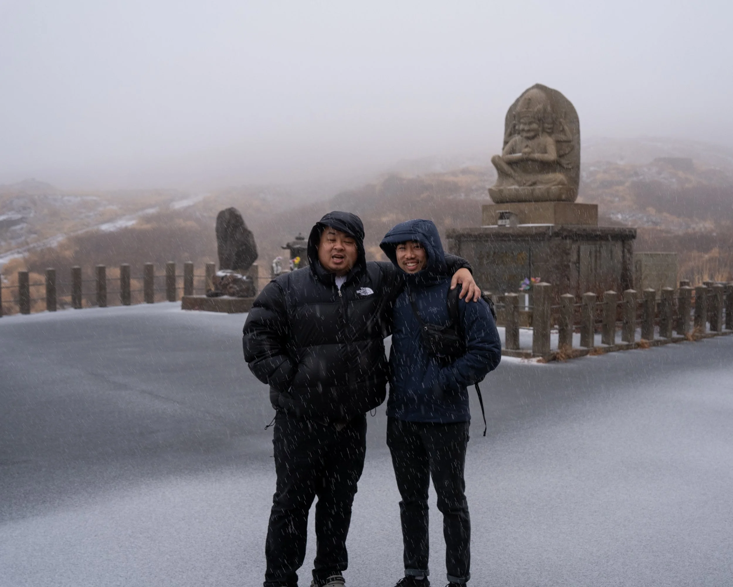 Two people wearing rain jackets standing in the rain with a mountain and a statue in the background.