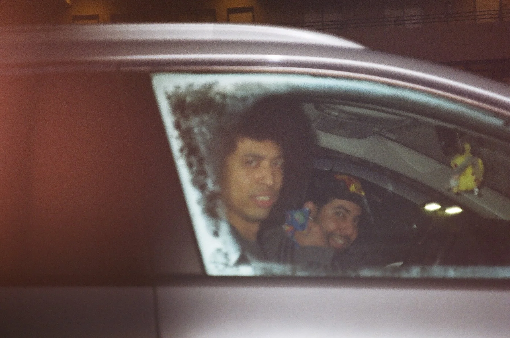 Two young men sitting in a car, taking a selfie in the rearview mirror, smiling and looking at the camera.