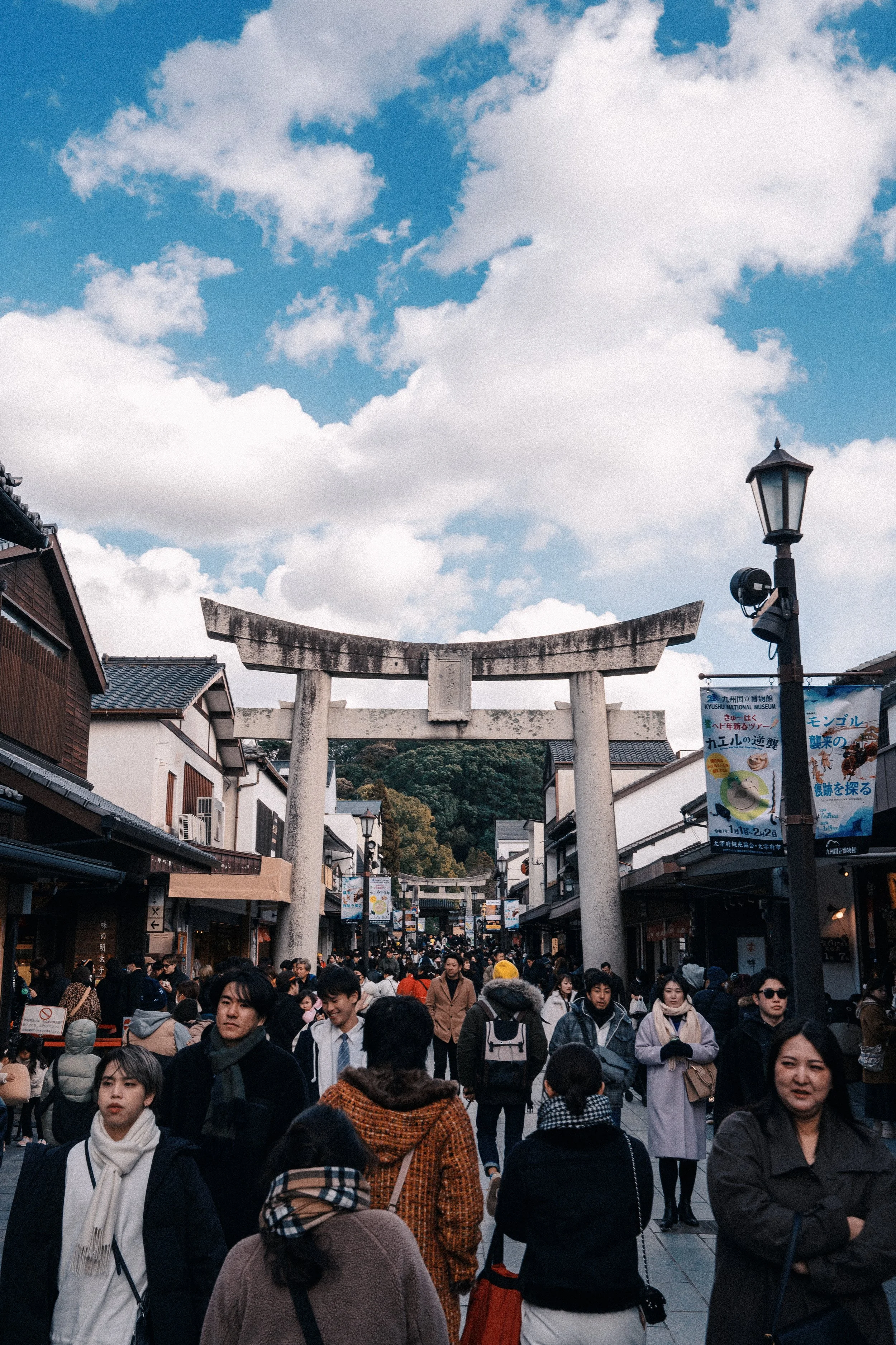 Crowd of people walking through an outdoor shopping street with traditional Japanese torii gate and shops on either side, under a cloudy sky.