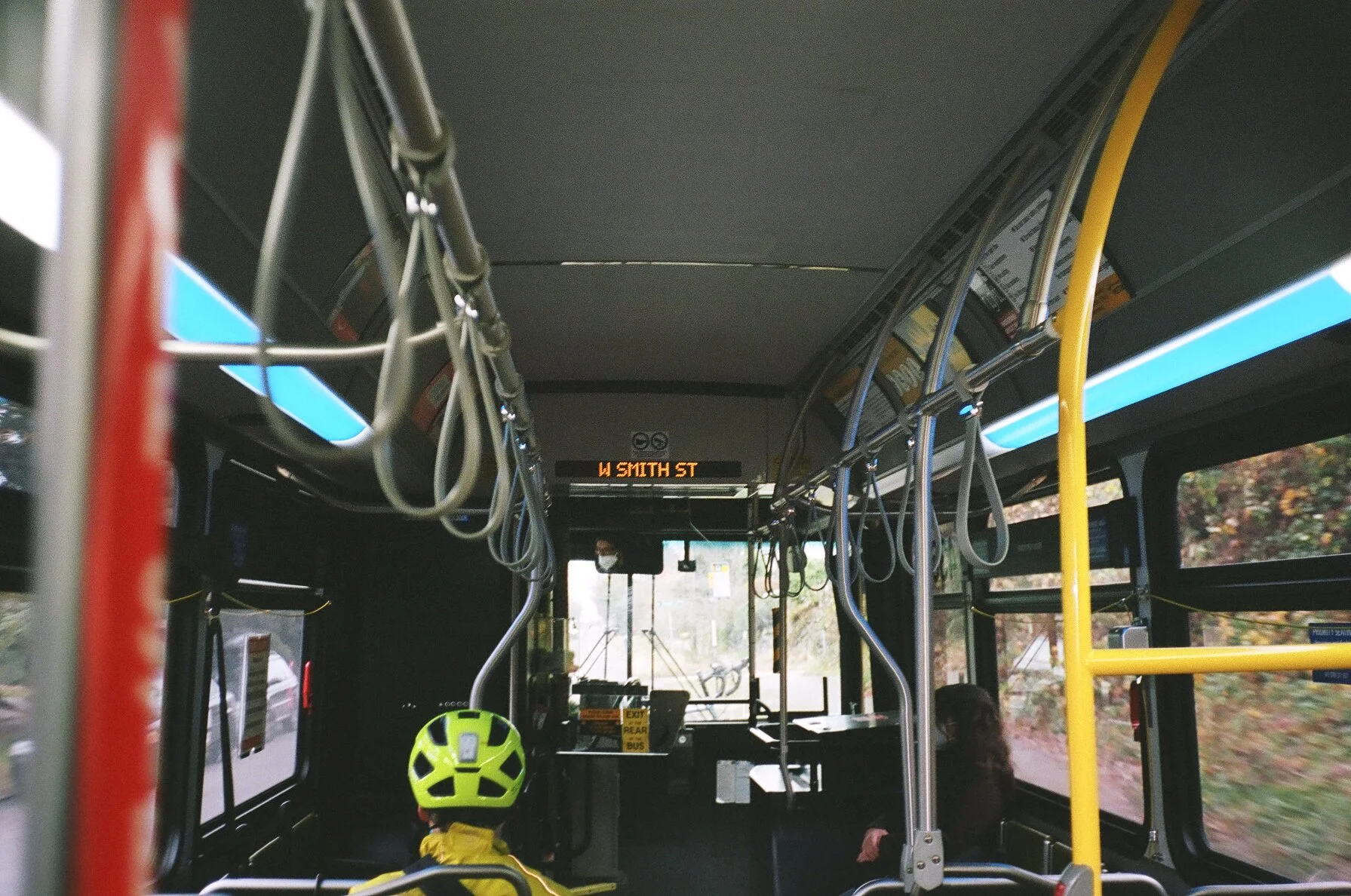 Inside of a city bus showing two passengers, including a person wearing a bright yellow bicycle helmet, and a digital display indicating the route to W Smith St.