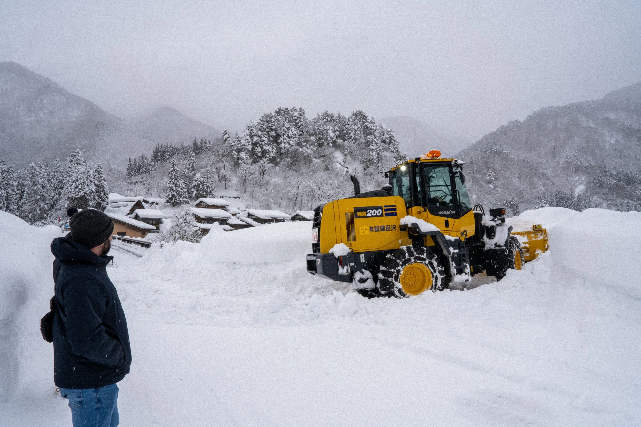 A person in a dark jacket and beanie watching a yellow snowplow push snow in a snowy mountain village with snow-covered trees and mountains in the background.