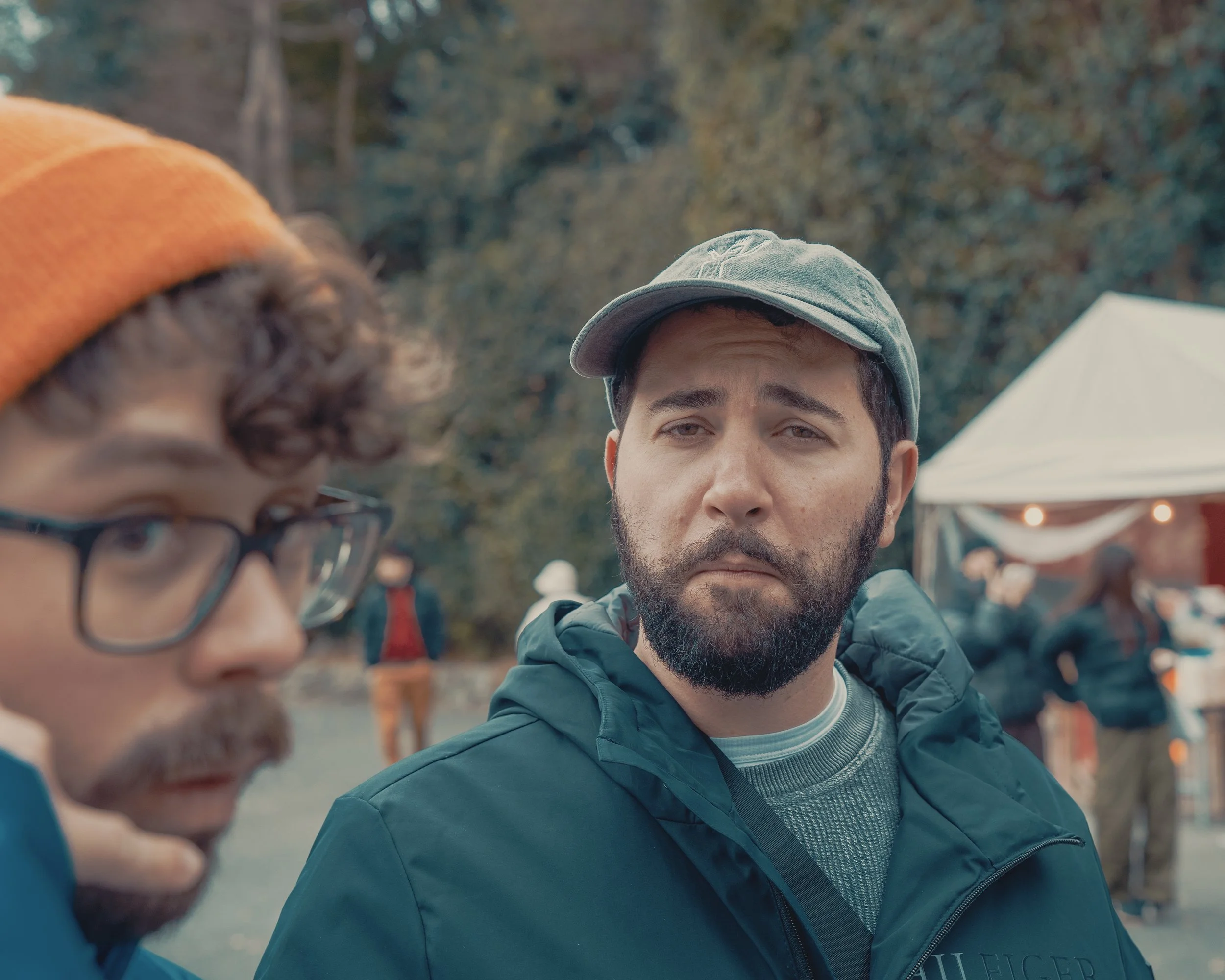 Two men outdoors at a gathering, with one man in the foreground and wearing glasses and an orange beanie, and the other in the background with a gray cap and a beard, looking serious. There is a white tent and other people in the blurred background.