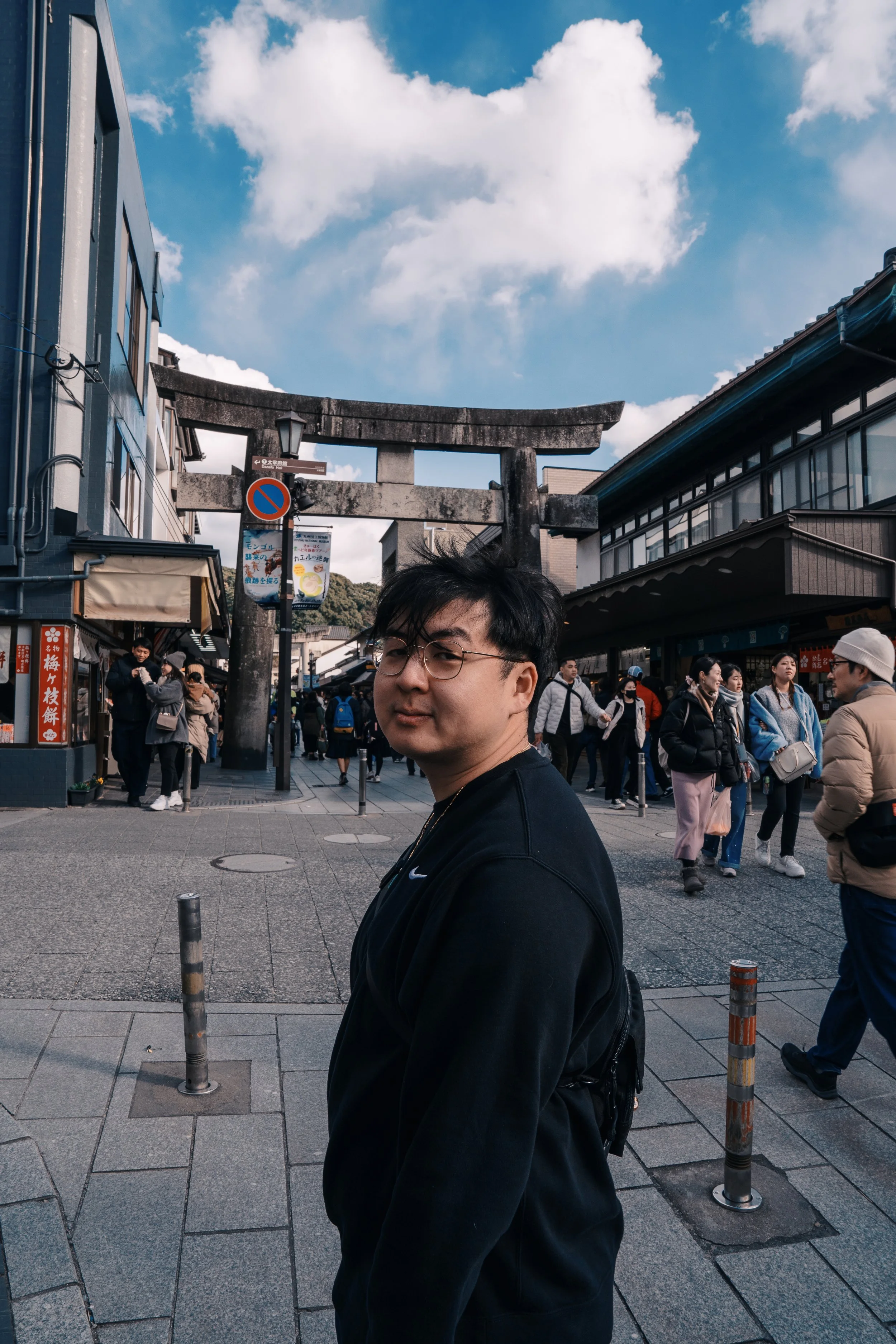 A young man with glasses and a black sweatshirt stands in front of a large traditional Japanese torii gate in a busy shopping street with many people, shops, and signs under a partly cloudy sky.