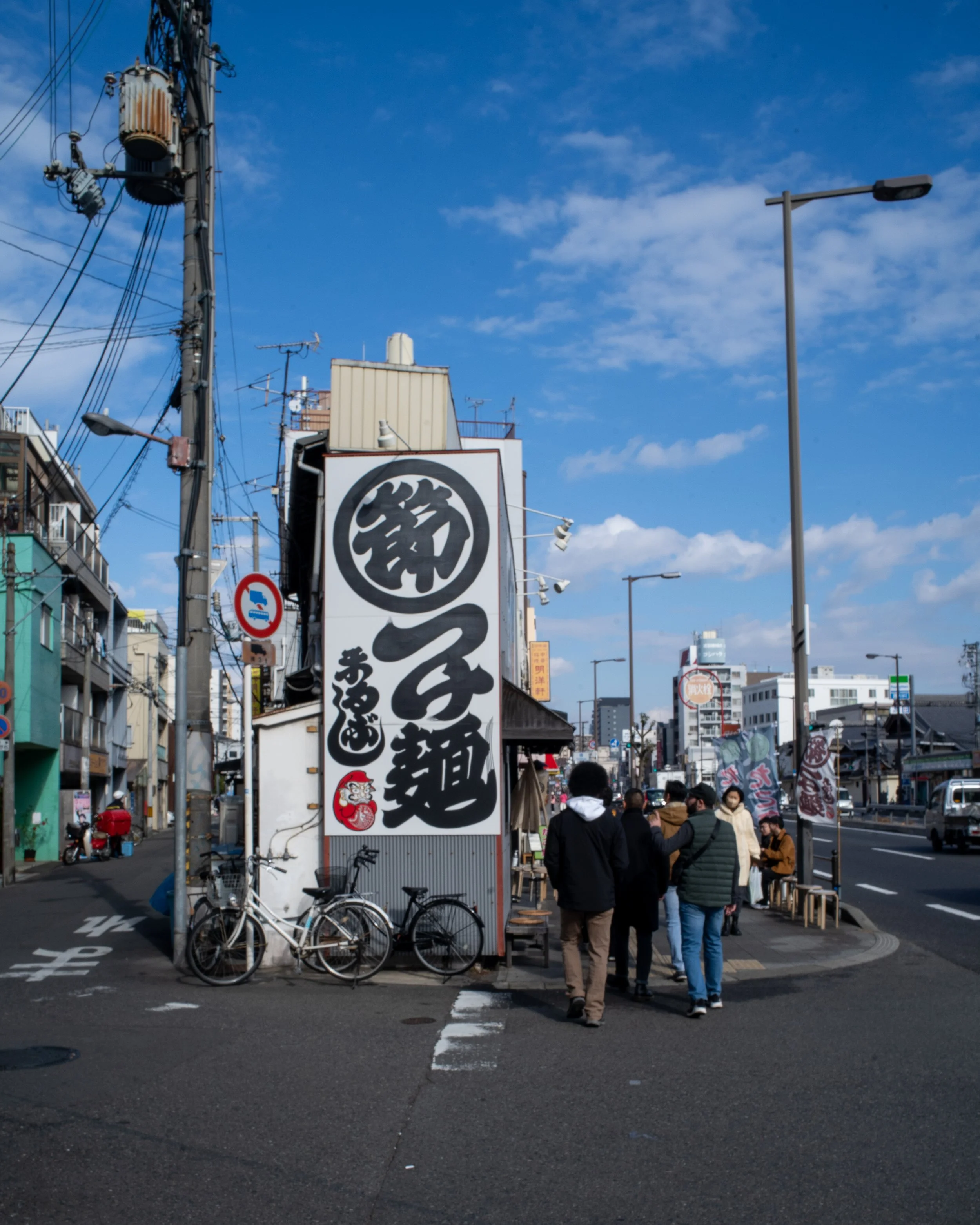 Street view in Japan with a large vertical sign featuring Japanese characters, bicycles parked nearby, and people walking along the sidewalk, under a blue sky with some clouds.