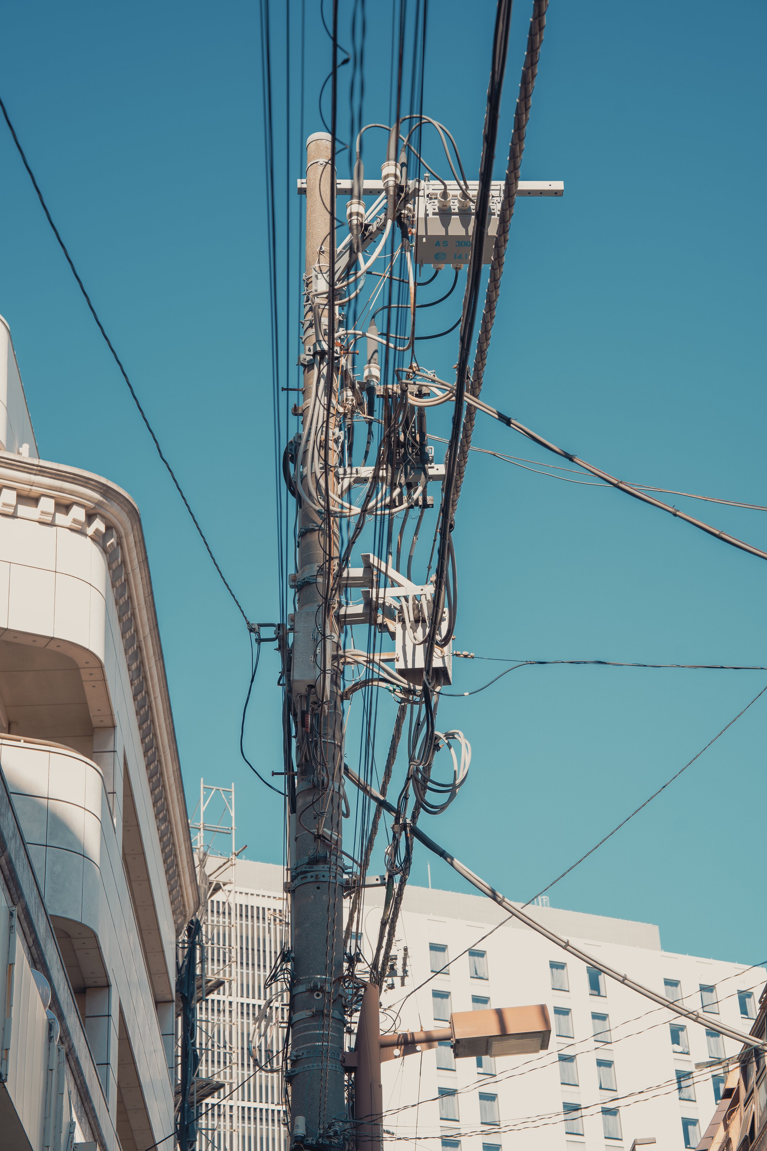 A utility pole with numerous electrical wires and equipment against a clear blue sky, with parts of surrounding buildings visible.