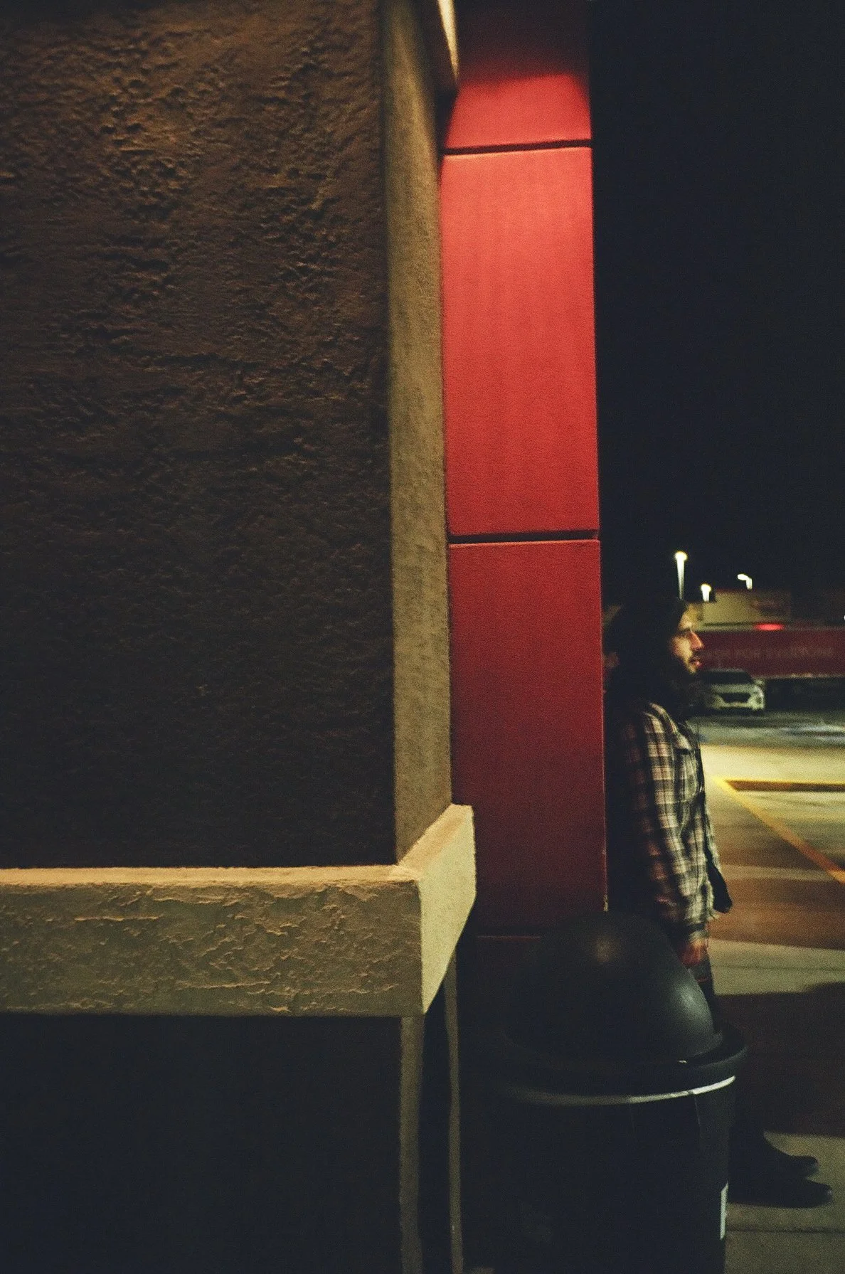 A person standing at night near a wall and red panel, with a trash can in front, in a parking lot with cars and lights in the background.