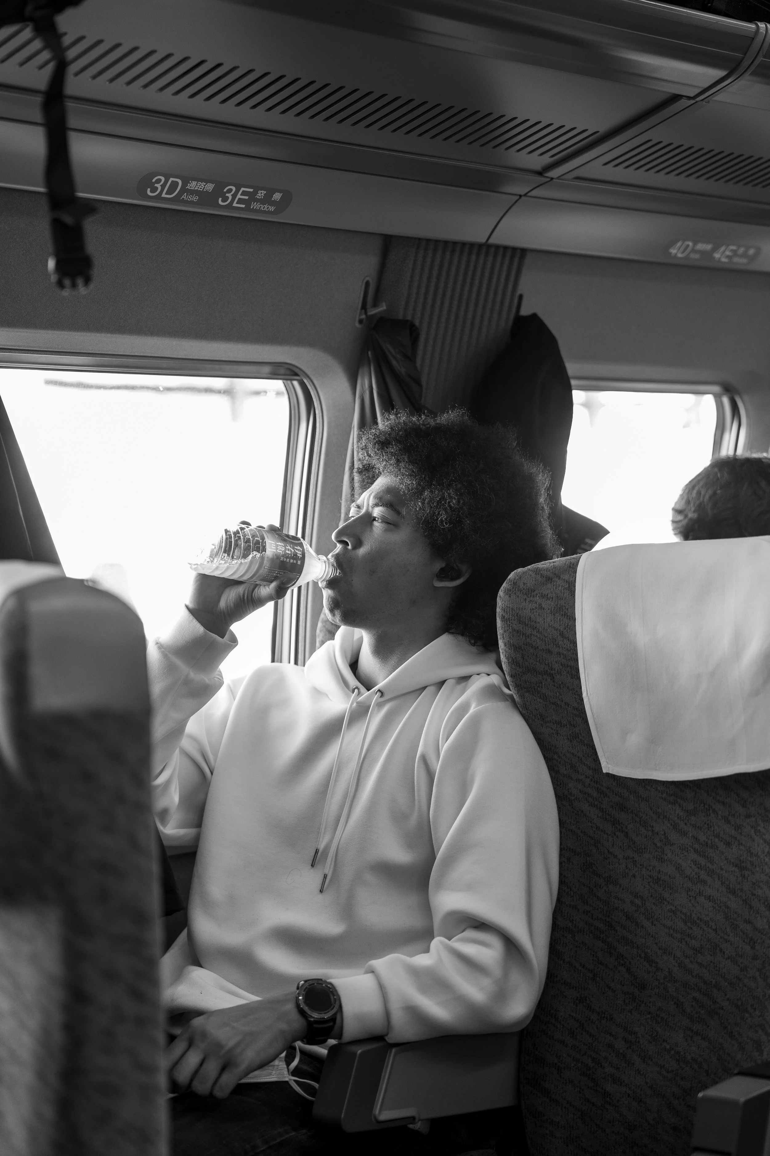 A young man with afro hair, wearing a white hoodie, is sitting on a train and drinking water from a plastic bottle.