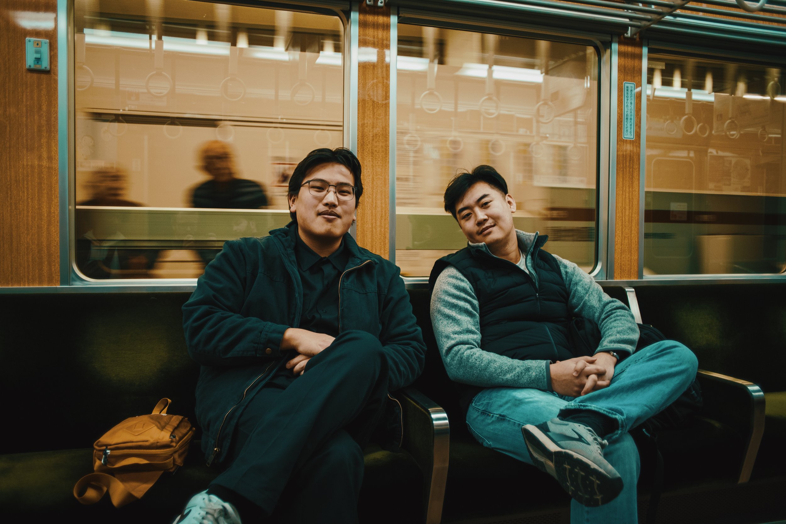 Two young men are sitting on a bench inside a train, looking at the camera. The train has wooden paneling and glass windows with reflections. There are blurred figures visible through the windows.