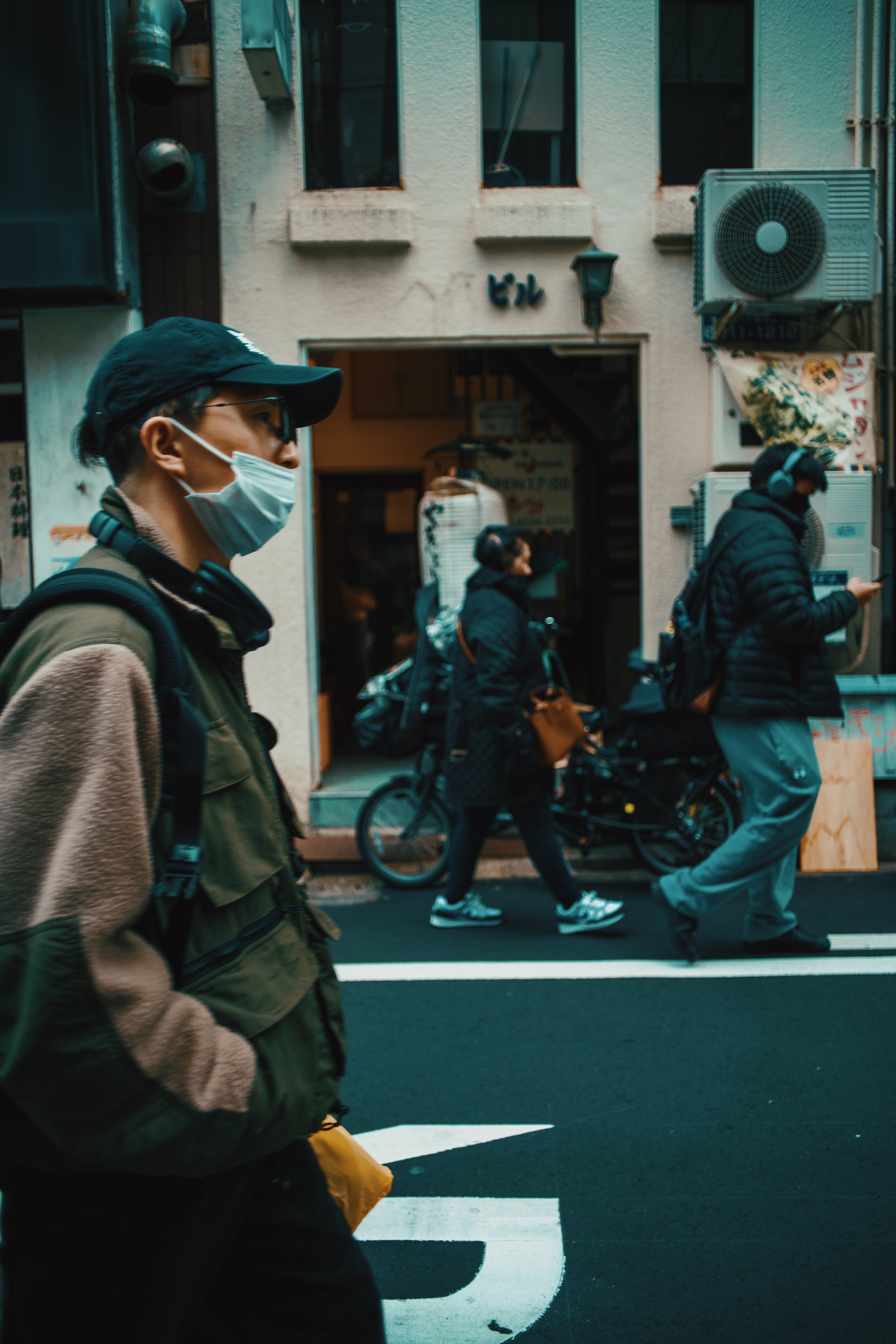 People walking on a city street, some wearing face masks and carrying backpacks, with buildings and a bicycle in the background.