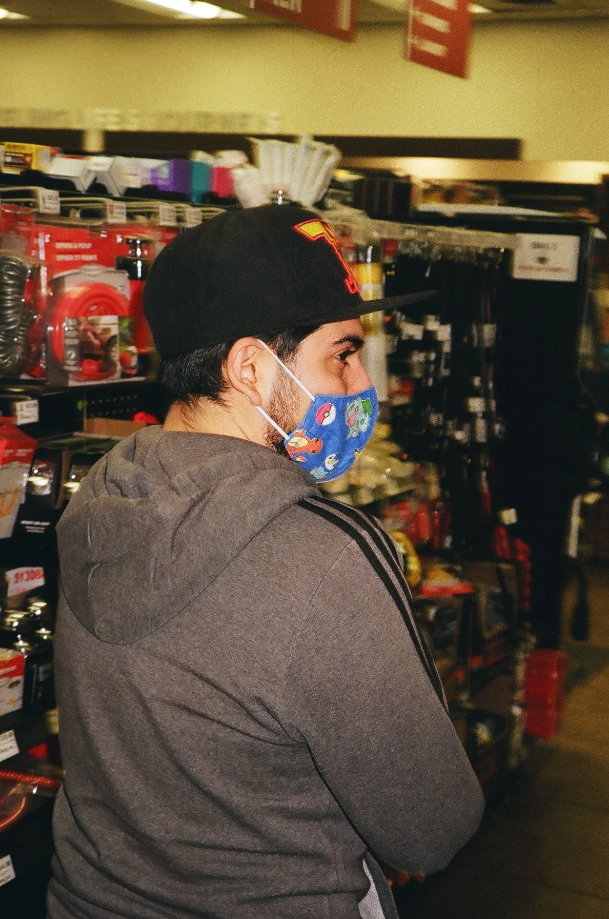 A man with a dark baseball cap, gray hoodie, and a colorful Pokémon-themed face mask shopping in a store aisle with various kitchen and household items on shelves.