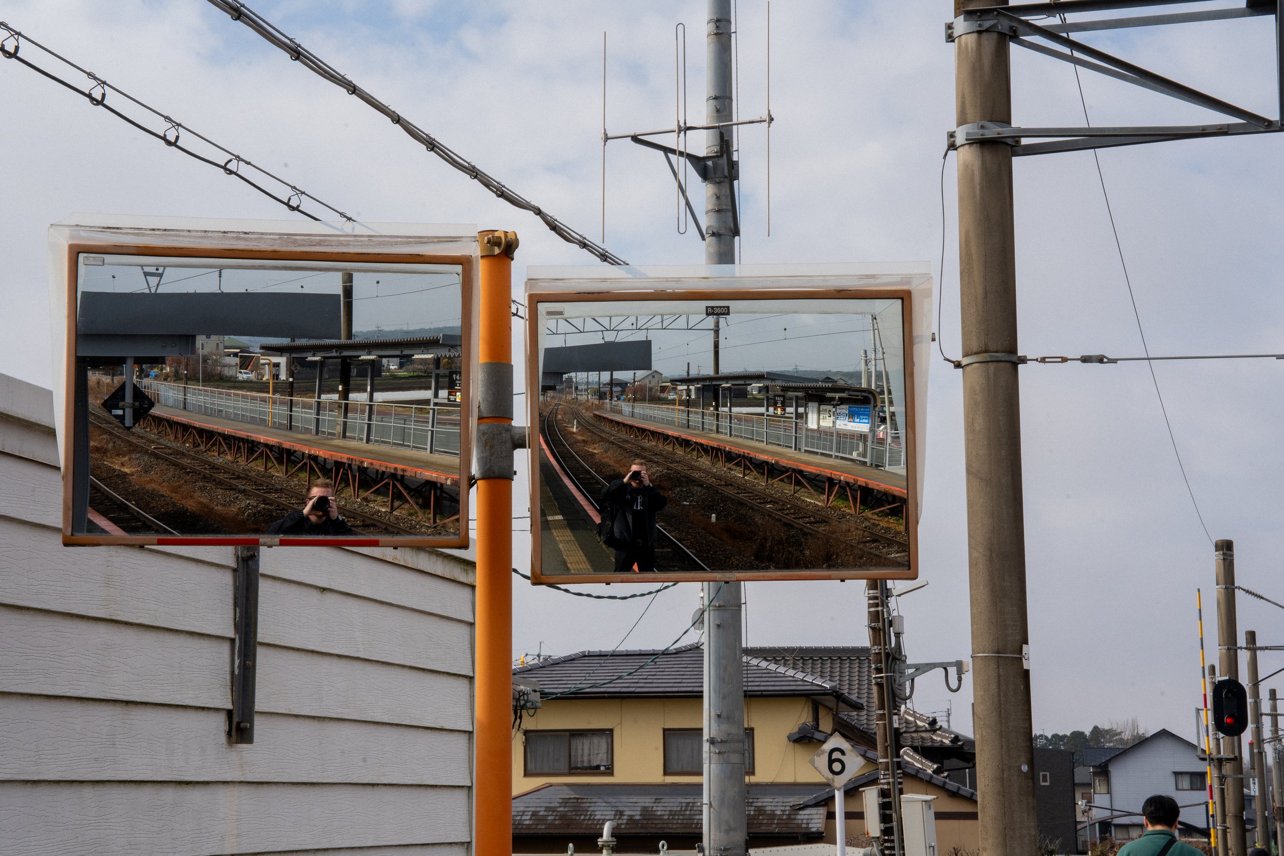 Two train station mirrors reflecting the platform, tracks, and a person taking a photo, with a suburban neighborhood in the background.
