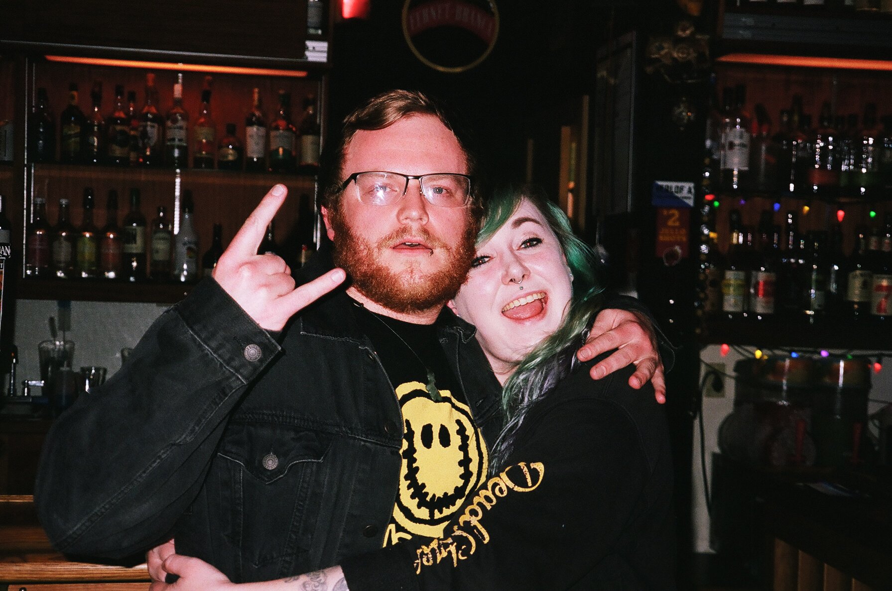 Two people hugging and smiling in a bar with liquor bottles on shelves behind them. The person on the left has glasses, a beard, and is making a rock sign with his hand. The person on the right has green hair and is sticking her tongue out.