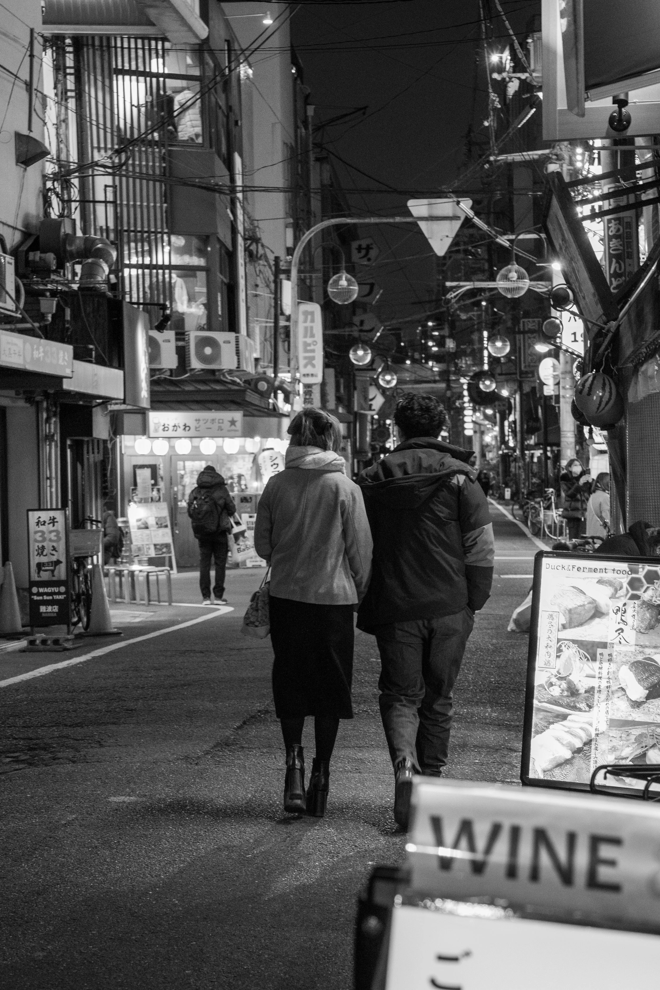 A black-and-white photo of a busy street at night with people walking. Two people are in the foreground, walking together, with the woman wearing a coat and boots and the man wearing a jacket. The street has illuminated signs, lanterns, and shopfront