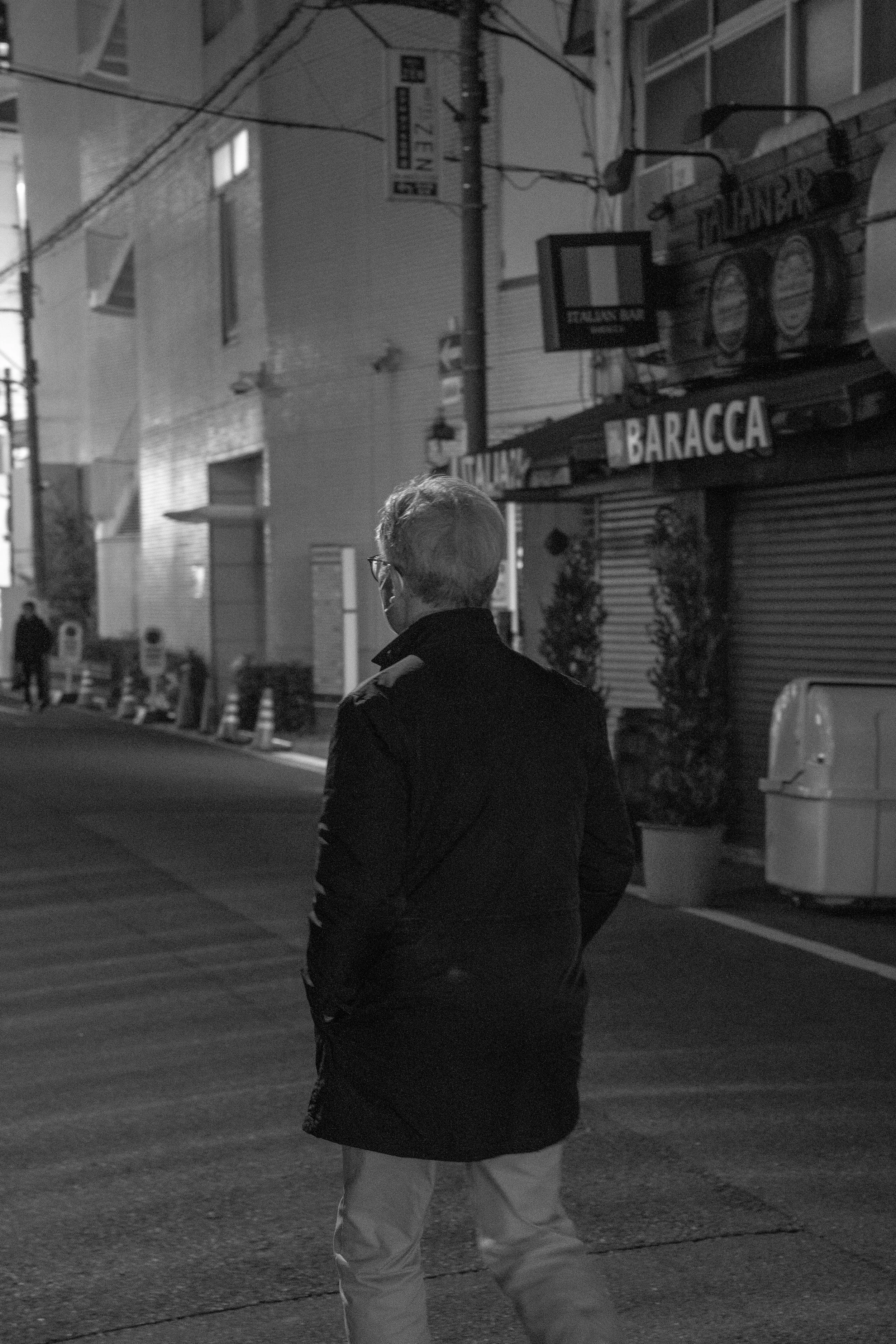 A man with glasses, wearing a black coat and light-colored pants, is walking on a city street at night. The scene is black and white, with buildings, streetlights, and closed storefronts visible in the background.