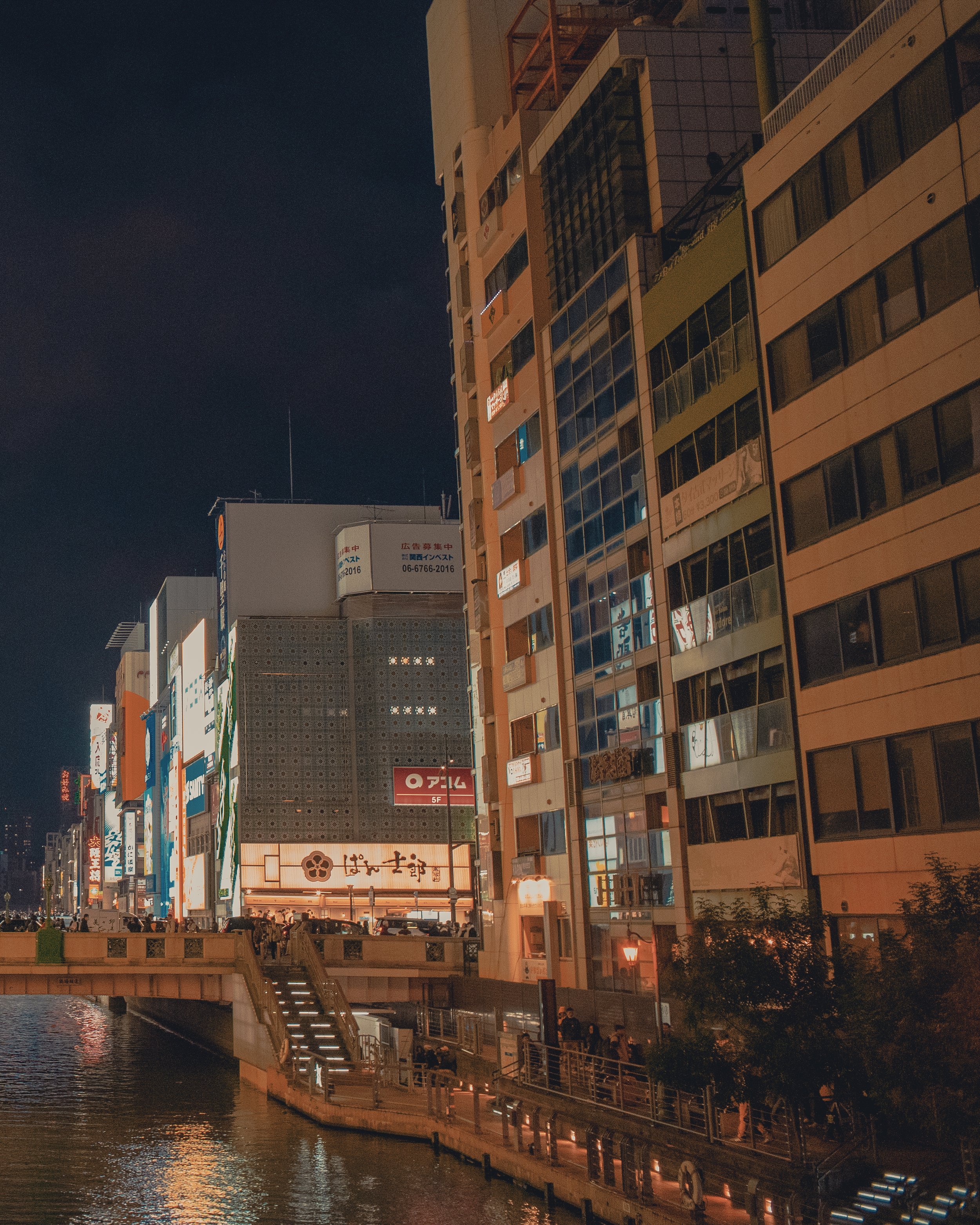 Night view of a cityscape with illuminated buildings and billboards along a canal, with a pedestrian walkway.