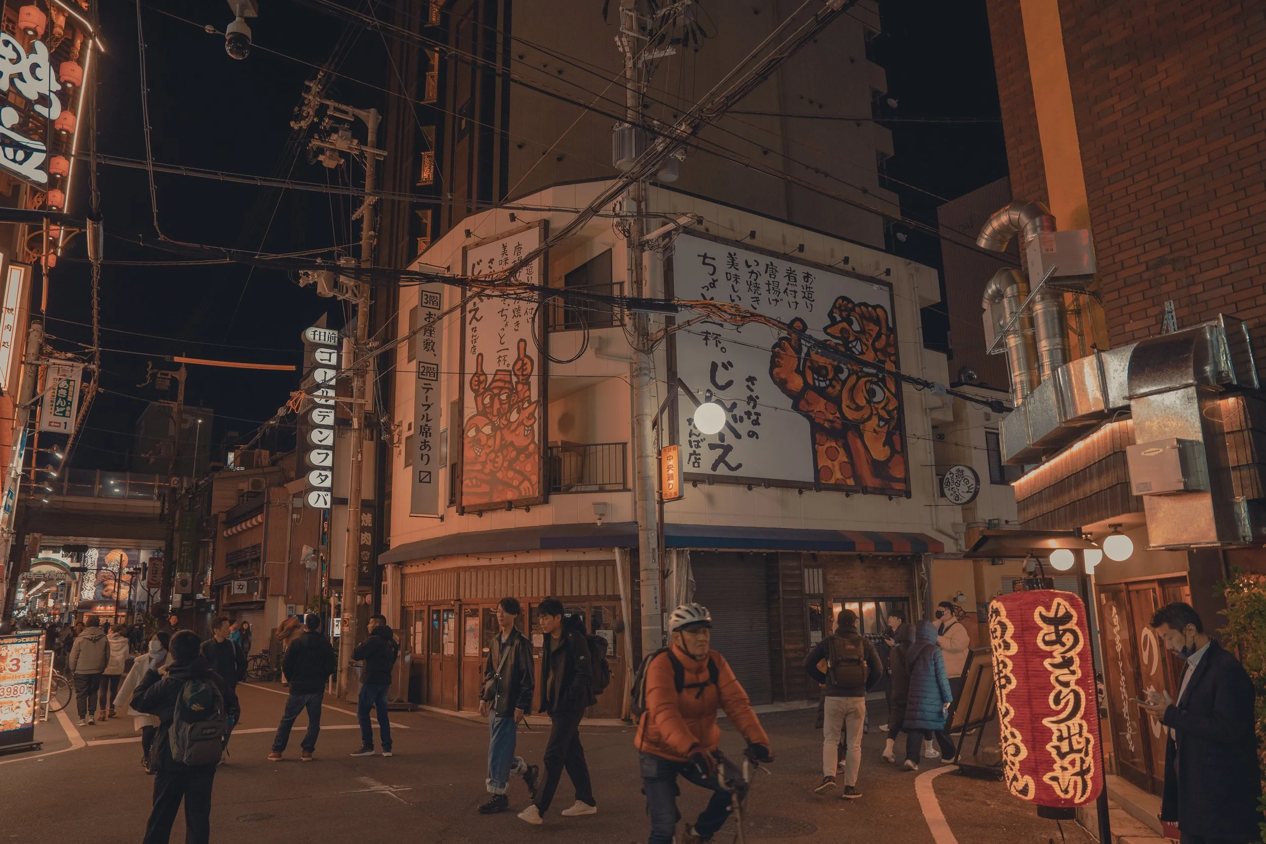 Night scene of a busy street corner in Japan with illuminated signs, banners, and people walking and cycling. Large mural of a tiger on the building, traditional lamp decorations, and various storefronts.
