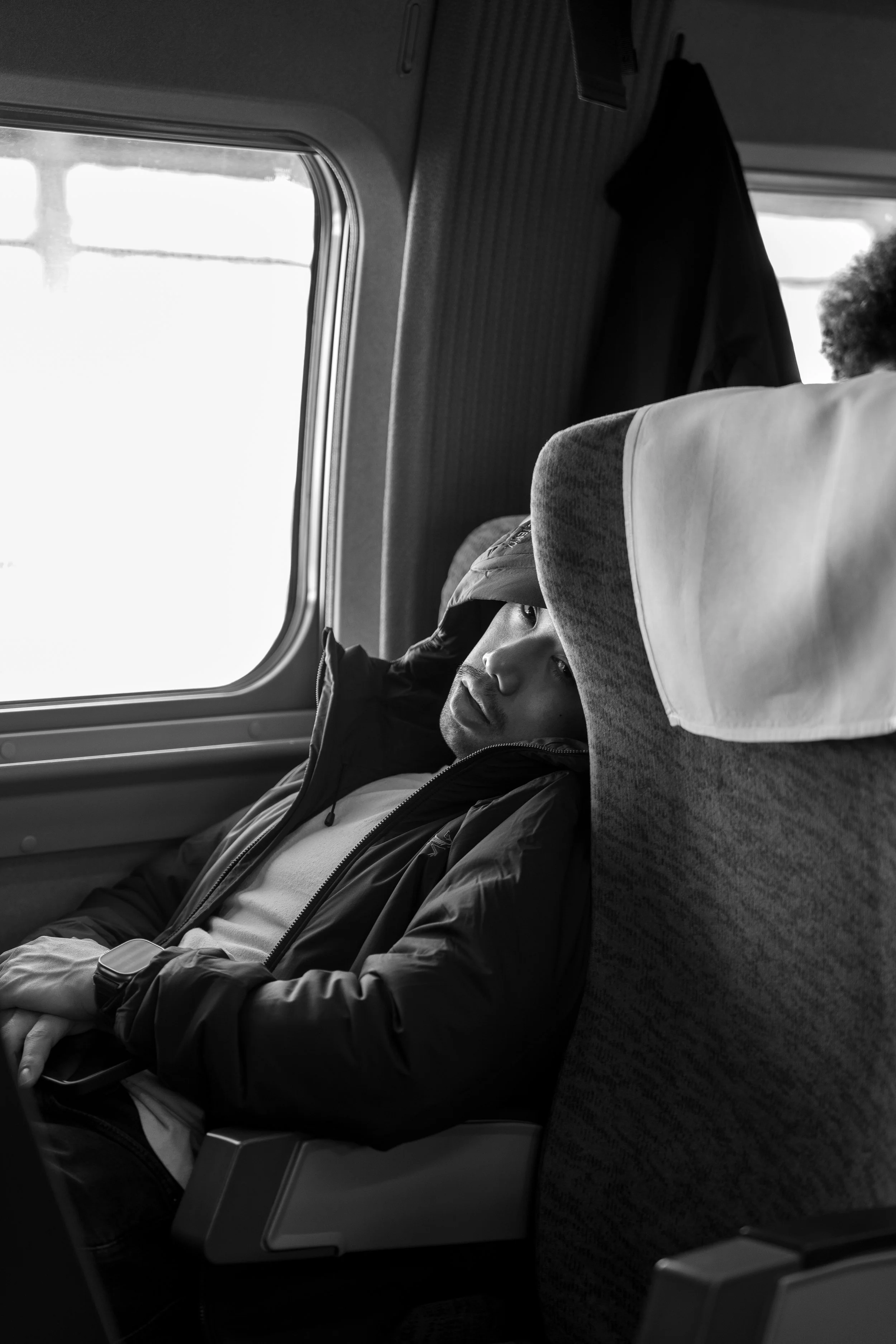 A person wearing a hooded jacket and a cap sitting on a train seat, resting with eyes closed, next to a window.