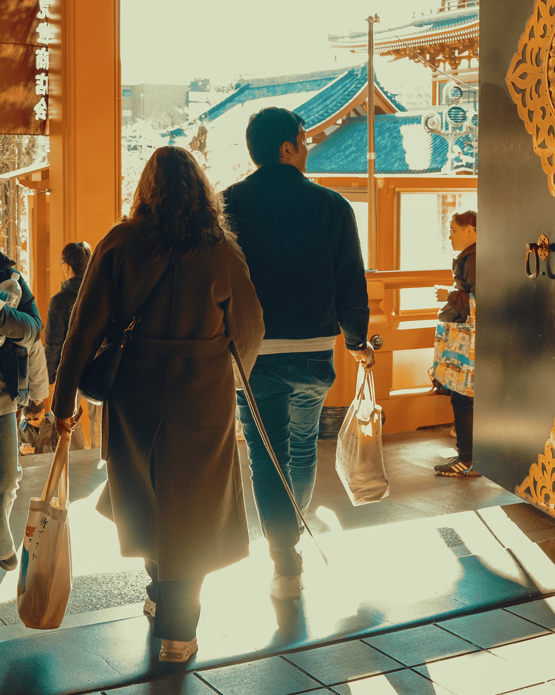 People entering a traditional Japanese shrine with blue tiled roof and wooden architecture, sunlight streaming in, and a woman with a shopping bag and a man carrying a tote bag.