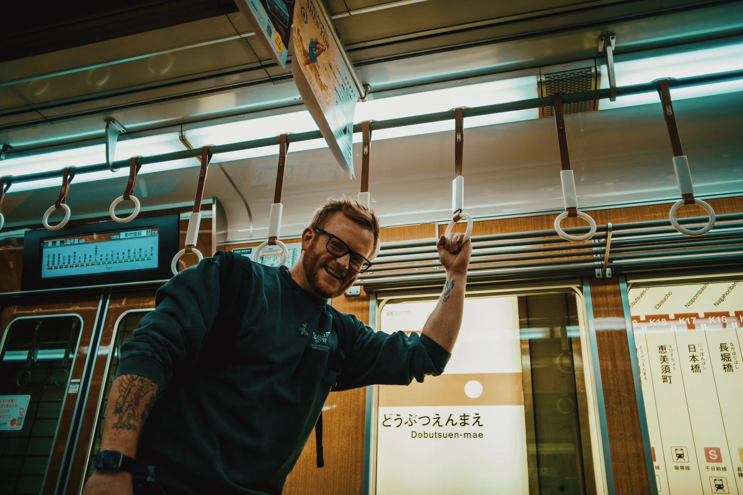 A man with glasses, tattoos, and a backpack smiling on a train, holding onto a hanging strap. The train has signs with Japanese characters and English translations, indicating a station name. The man appears happy and is standing near the doors of th