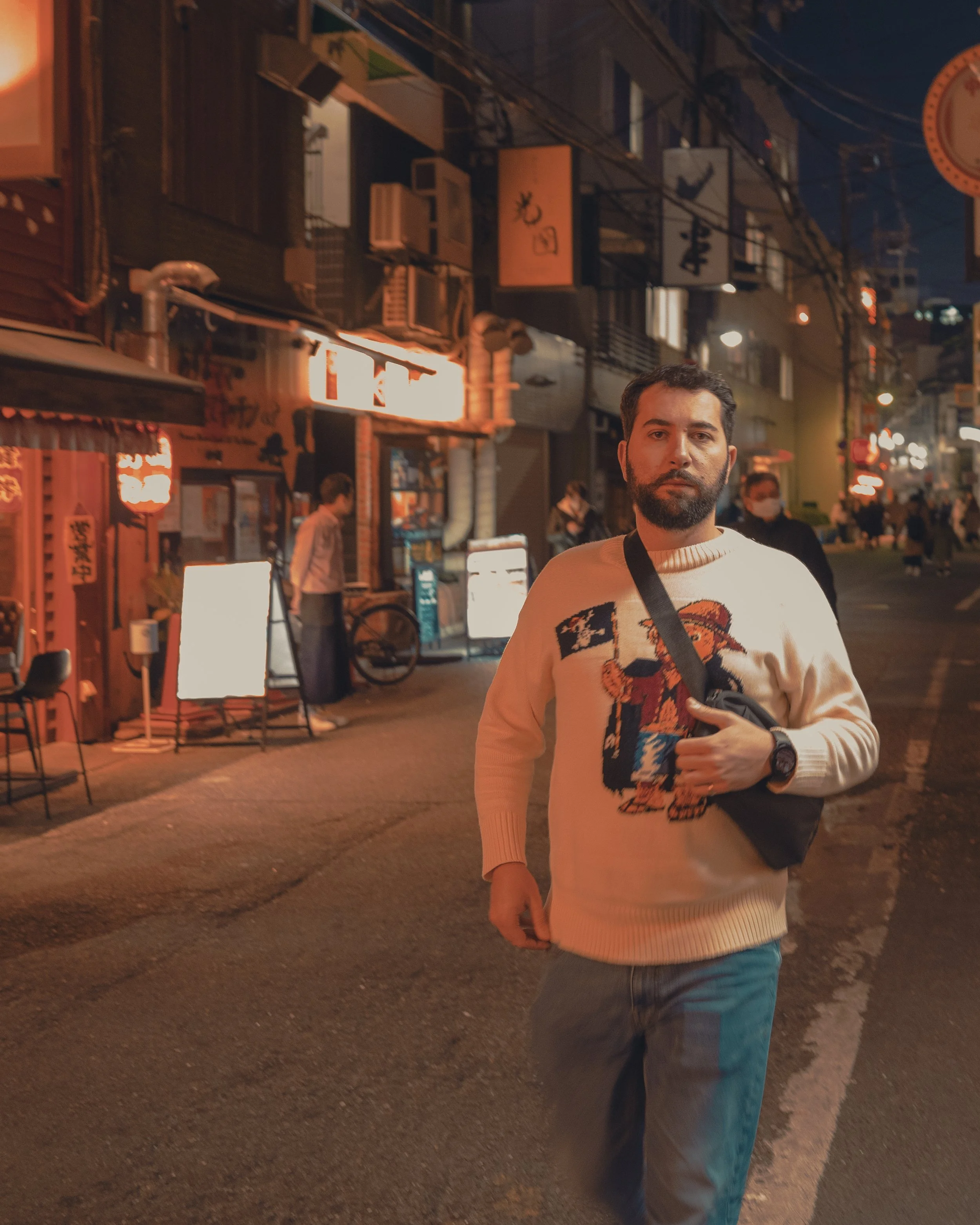 A man with a beard wearing a beige sweater with a graphic print, jeans, and a wristwatch stands on a city street at night. He has a camera strap across his chest and holds a small bag. The background shows illuminated signs, storefronts, other pedest