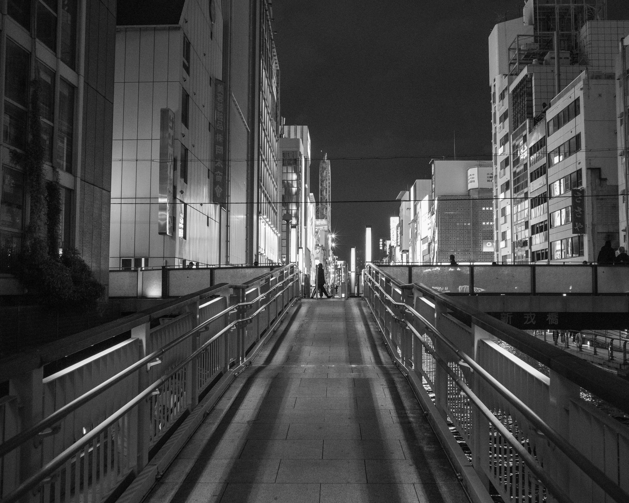 Night view of a city street in black and white, featuring a pedestrian bridge with a person walking on it, surrounded by tall illuminated buildings.