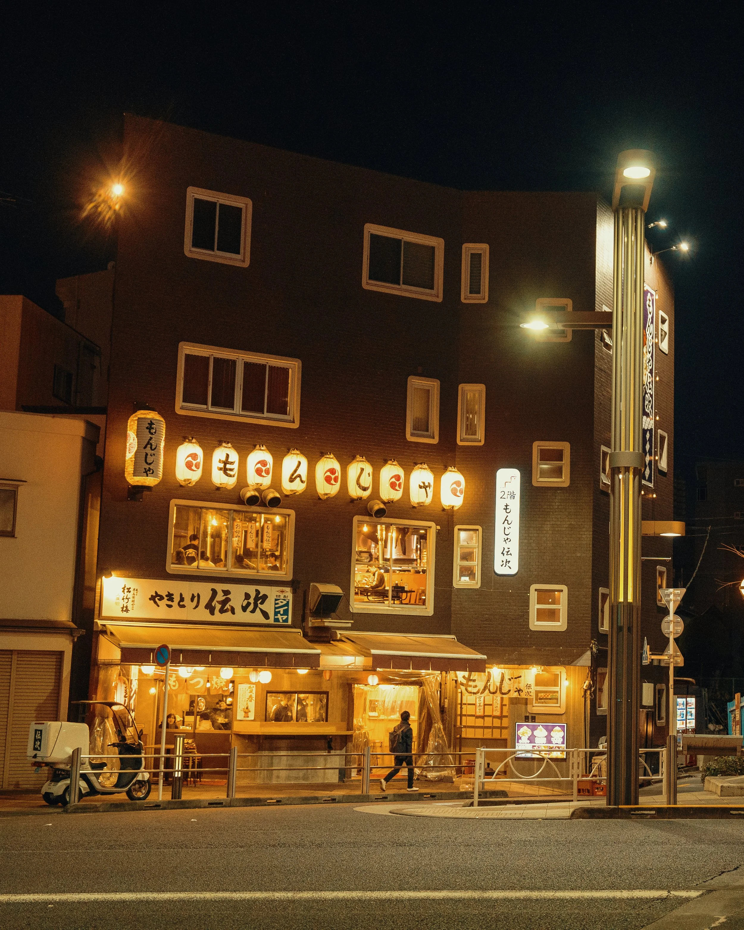Nighttime street view of a Japanese restaurant with illuminated paper lanterns and traditional signage, with a person walking by and a motorized scooter parked outside.