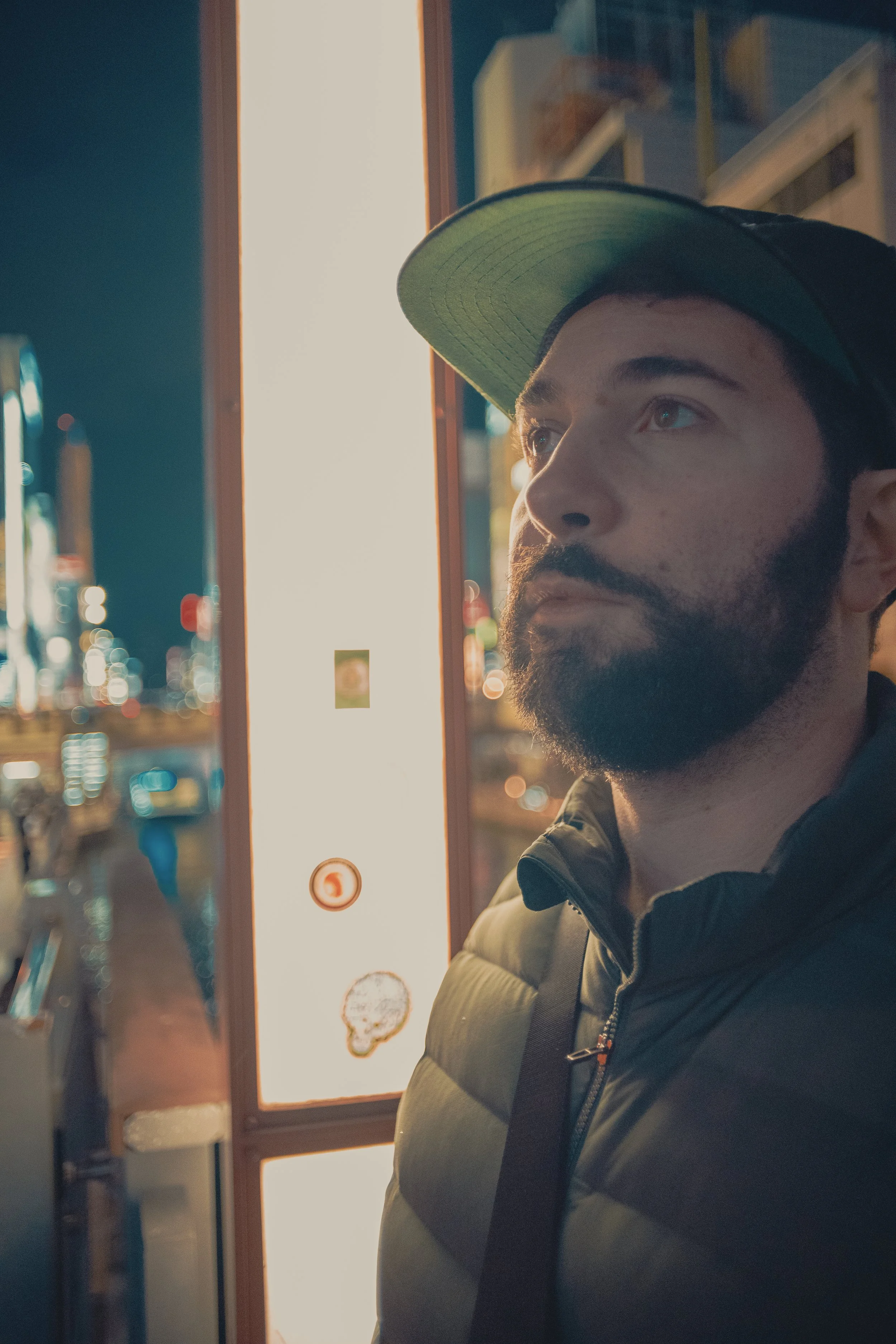 A man with a beard and light blue eyes wearing a black jacket and a green cap stands outside at night in an urban setting, looking thoughtfully into the distance.